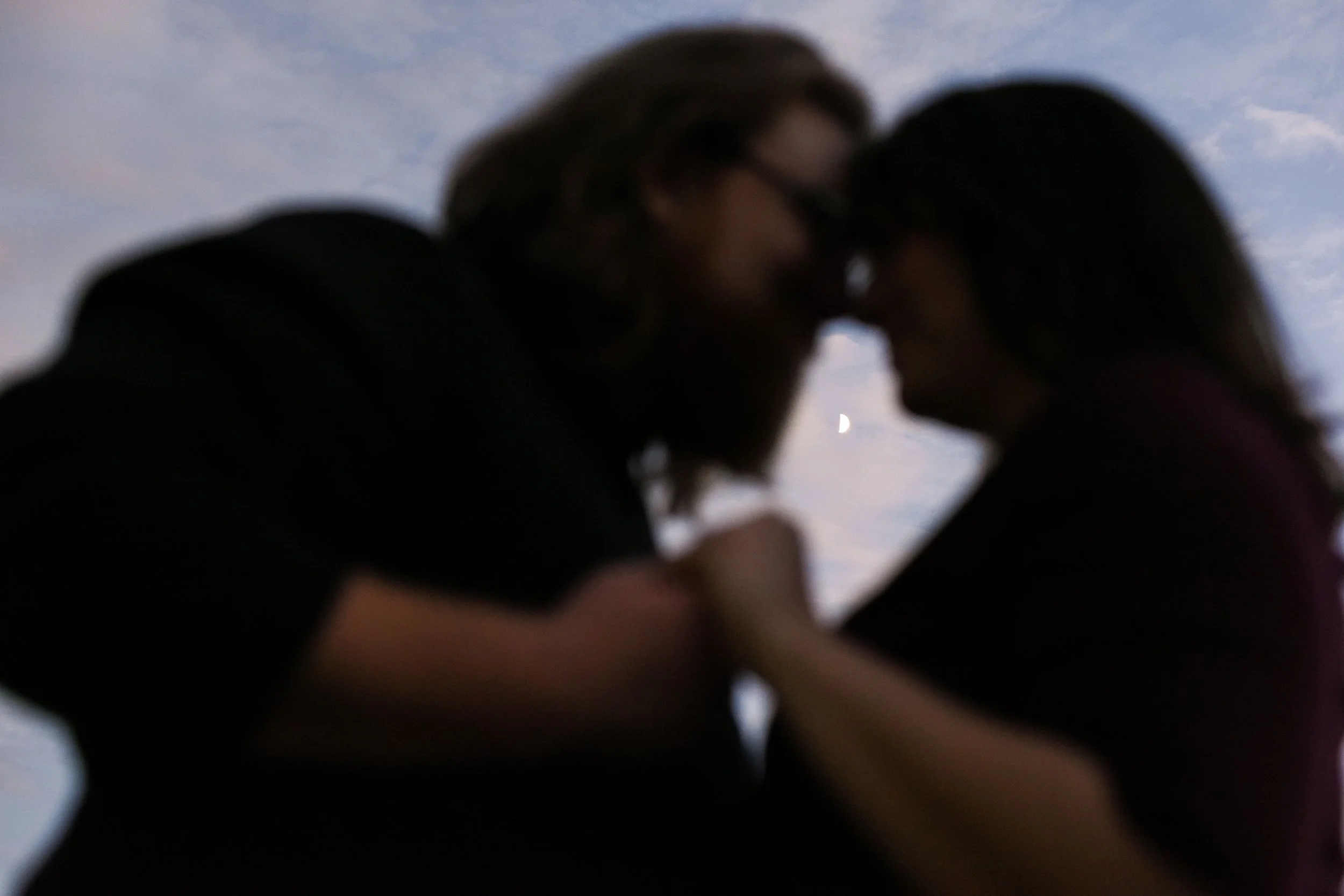 In the foreground, a couple stands holding hands with their foreheads pressed together, out of focus. In the background, the sky just after sunset with a half moon in focus in between their faces.