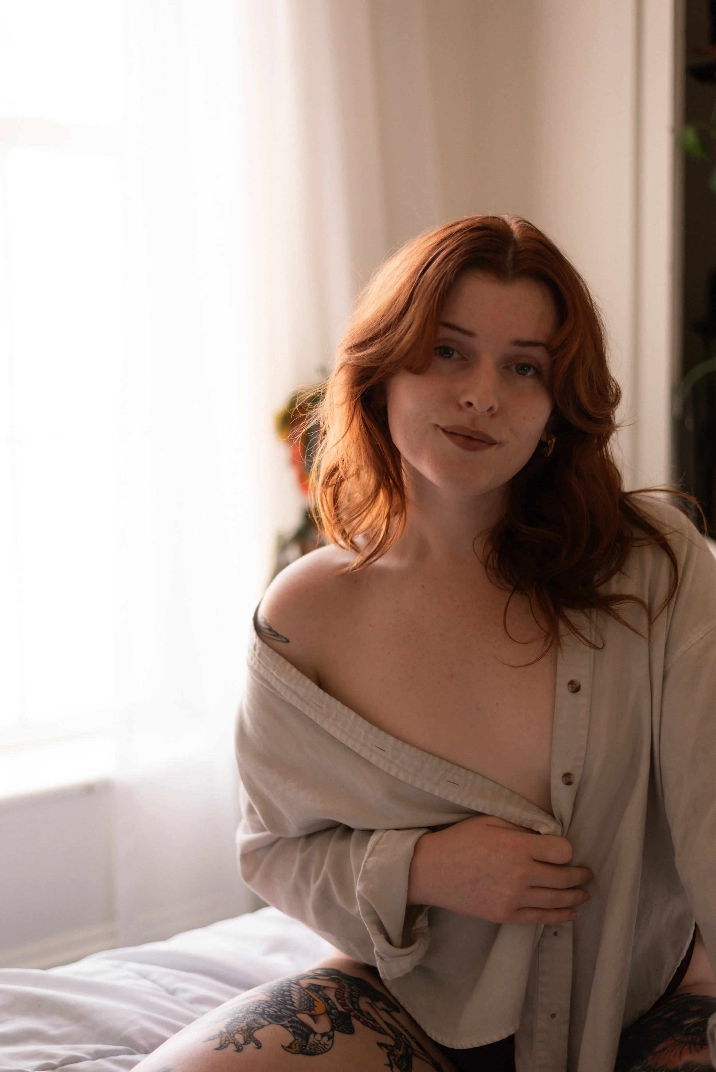 red haired woman with no makeup sits on a bed in front of a window looking directly into the camera while holding her button-up shirt closed at her chest