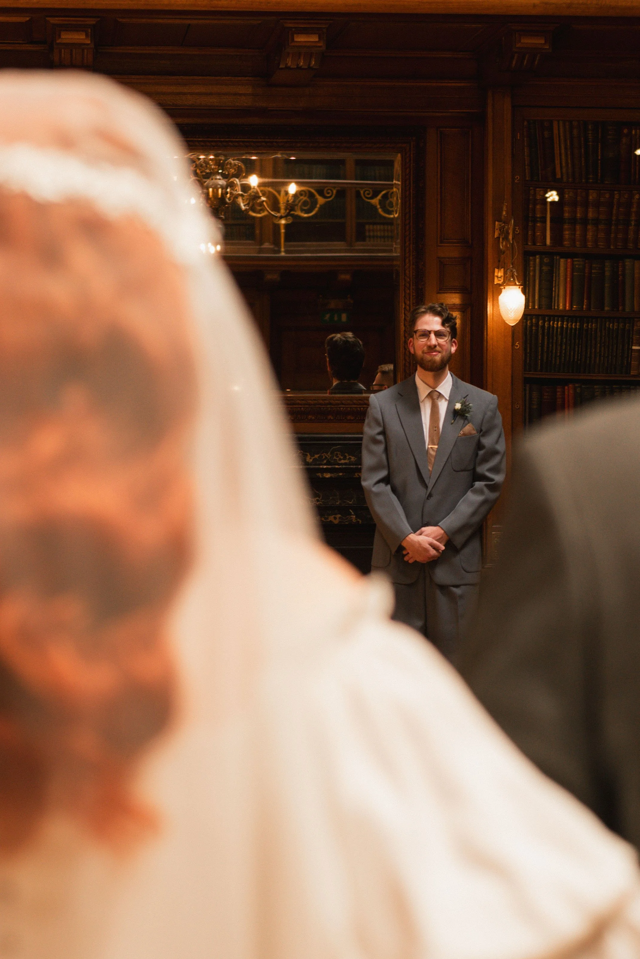 The image is over the shoulder of the bride and her dad as she walks down the aisle towards her groom, who stands in the center of the photo in focus smiling at his bride with his hands clasped.
