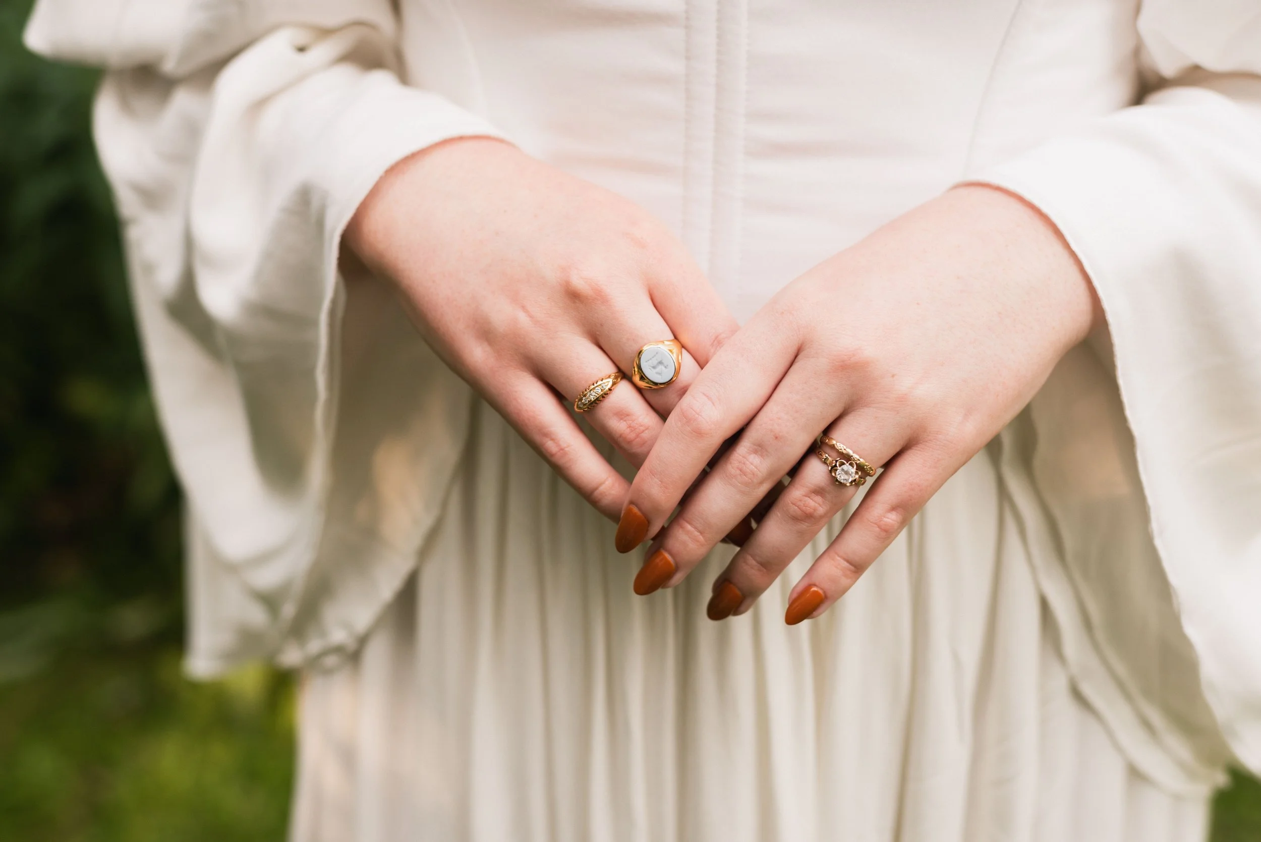 a bride stands with her hands in front of her, and this is a close up shot of her rings. You can see her engagement ring, new wedding band, light blue cameo ring, and other gold jewelry.