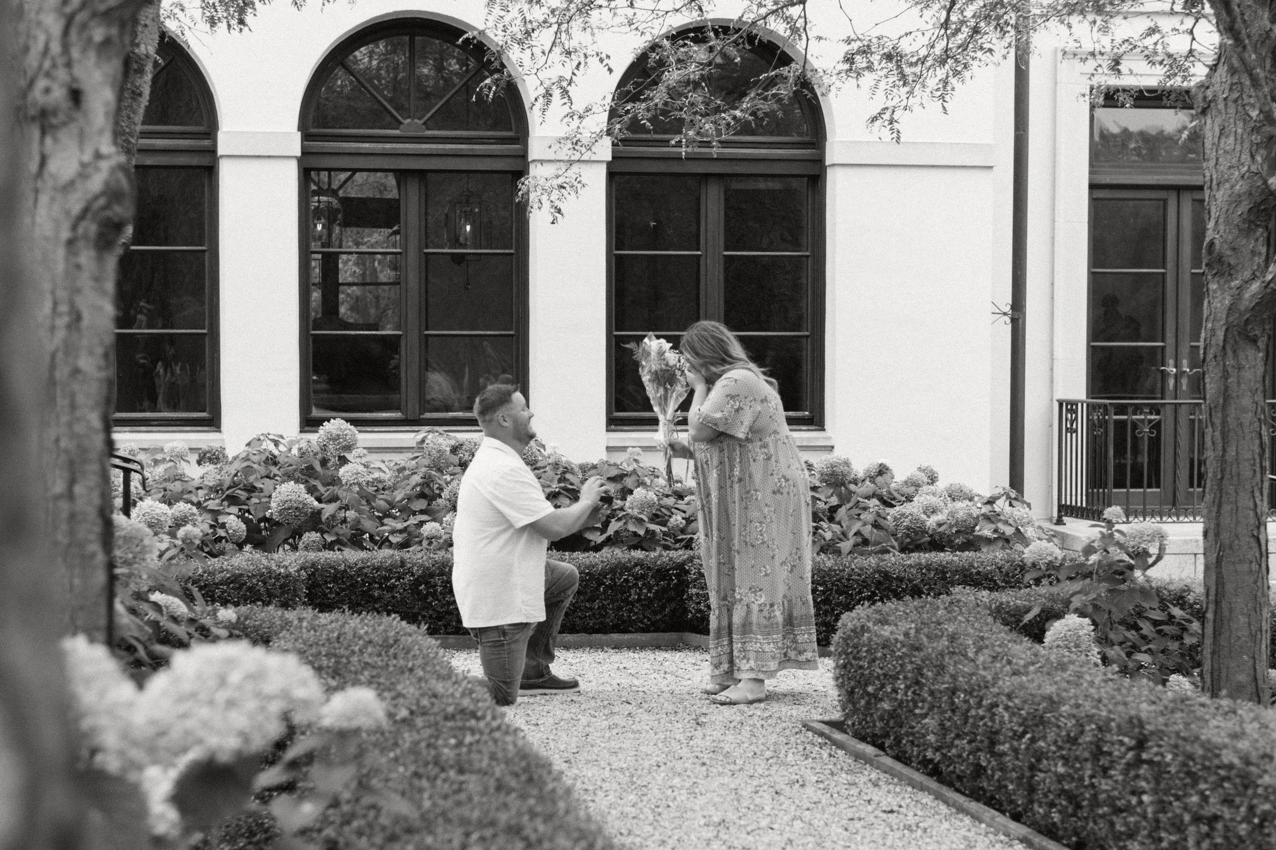 black and white image taken in a garden of a man on one knee holding a ring box in front of a standing woman in a long dress, with one hand over her mouth in surprise (she said yes to the proposal, by the way)