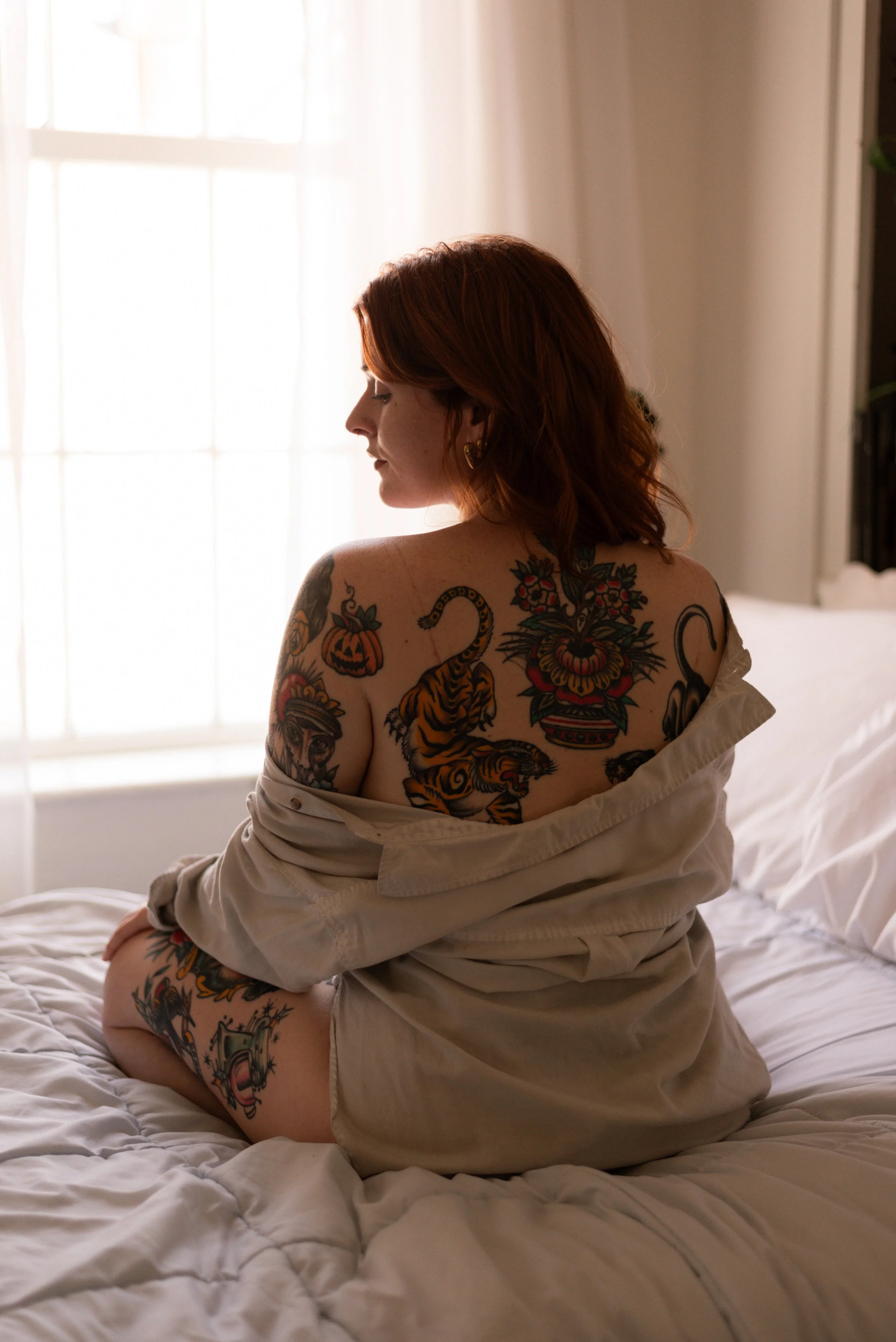a red haired woman with tattoos sits on a bed in front of a window facing away from the camera. She's looking down her left shoulder and her button up shirt is draped off of her shoulders showing her american traditional style tattoos