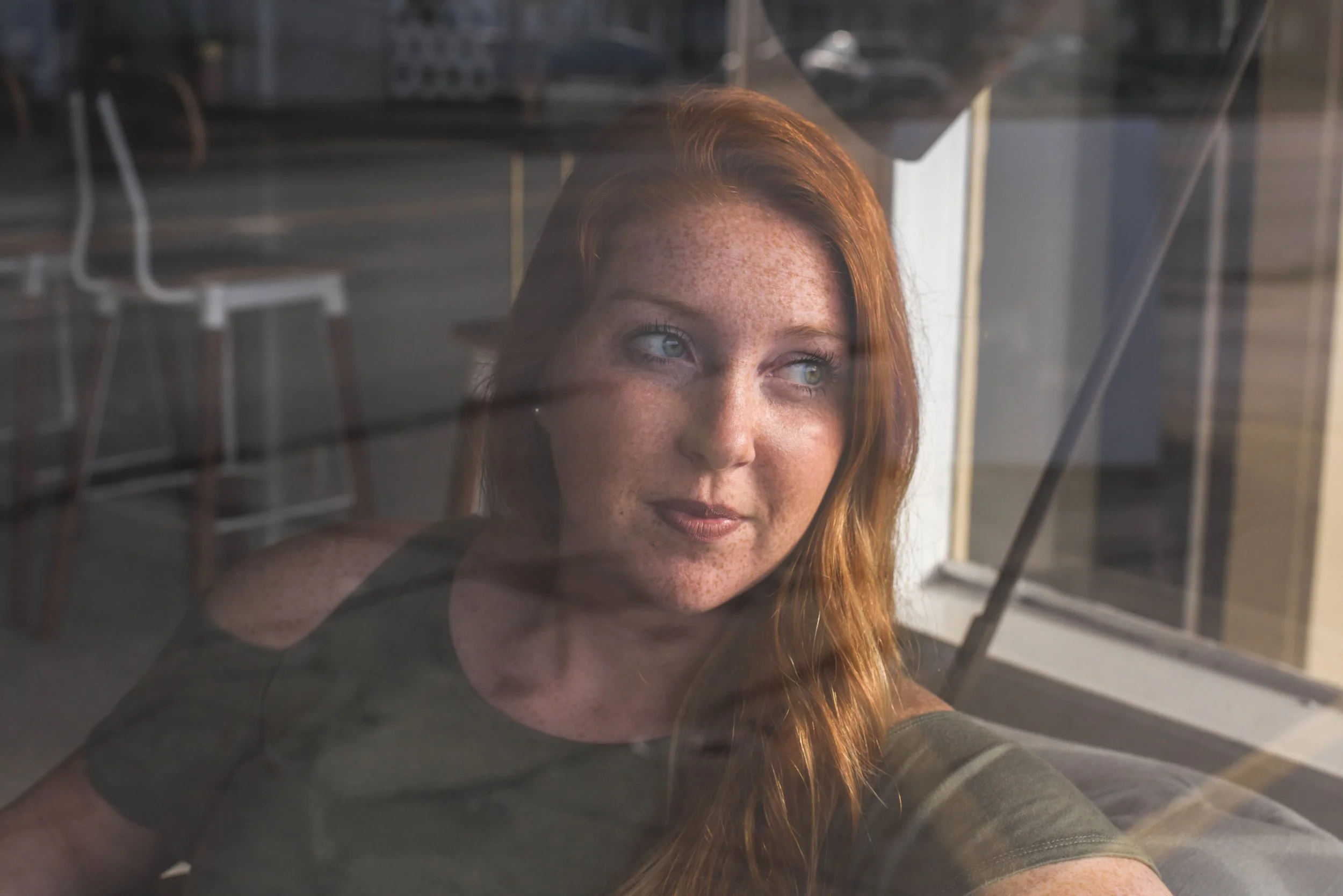 Red headed woman in an olive green shirt sitting in a coffee shop, looking through a glass window