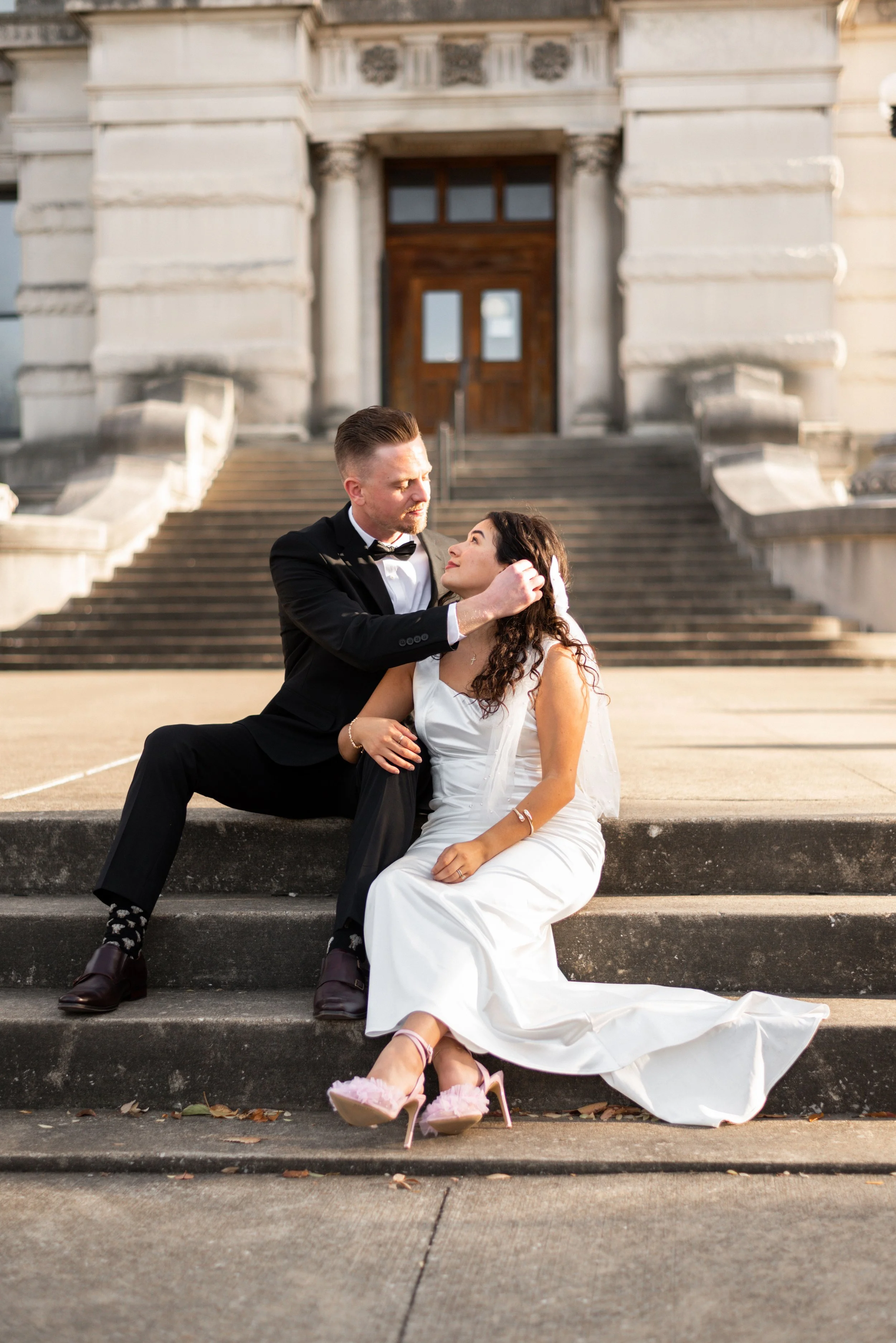 a bride and groom sit on the steps outside the Old Vanderburgh County Courthouse in downtown Evansville, Indiana. He is wearing a black suit and she is wearing a white satin dress with pink high heels, and they're gazing at each other.