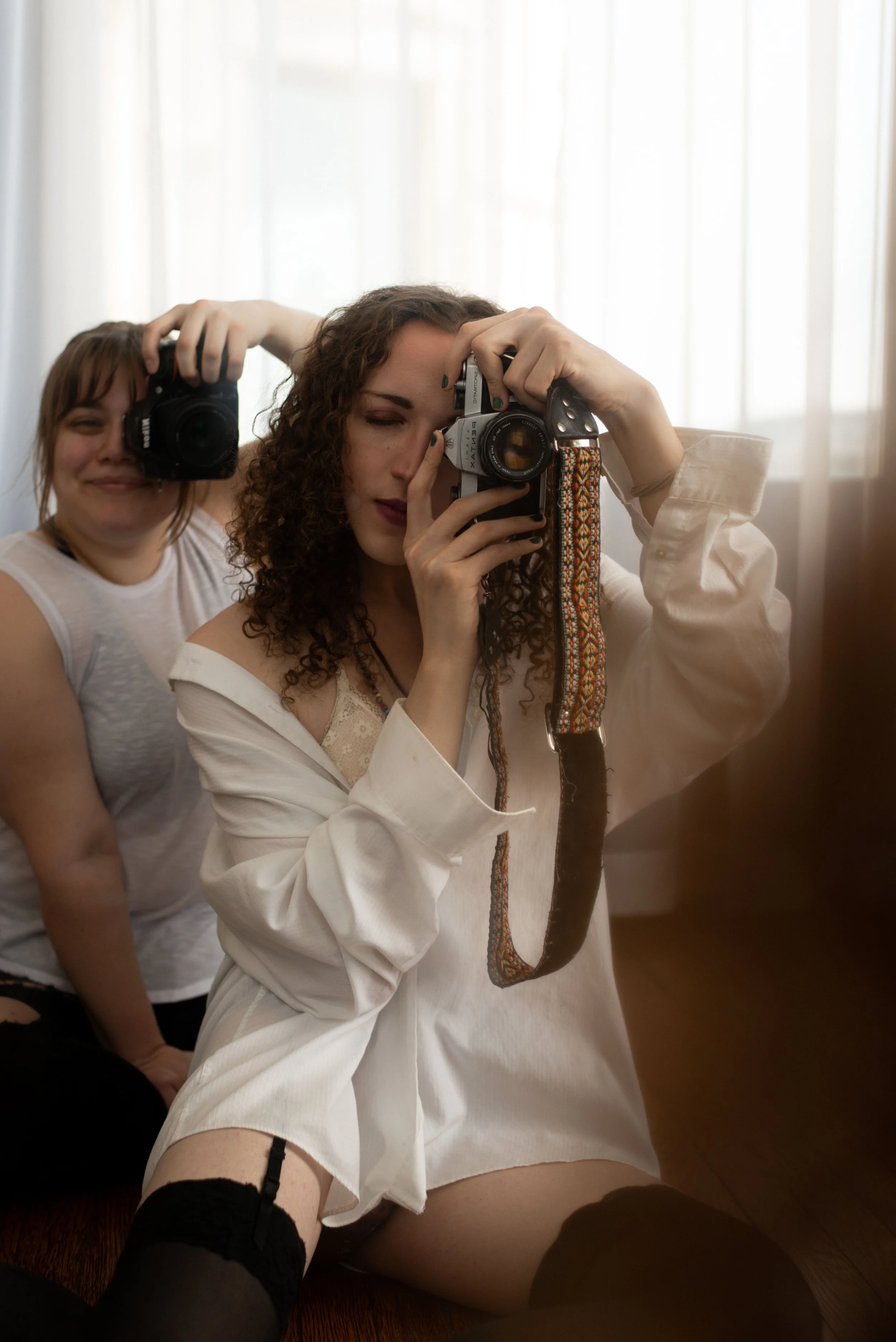 two women are seated next to each other, both with cameras in hand, taking a photo in a mirror.