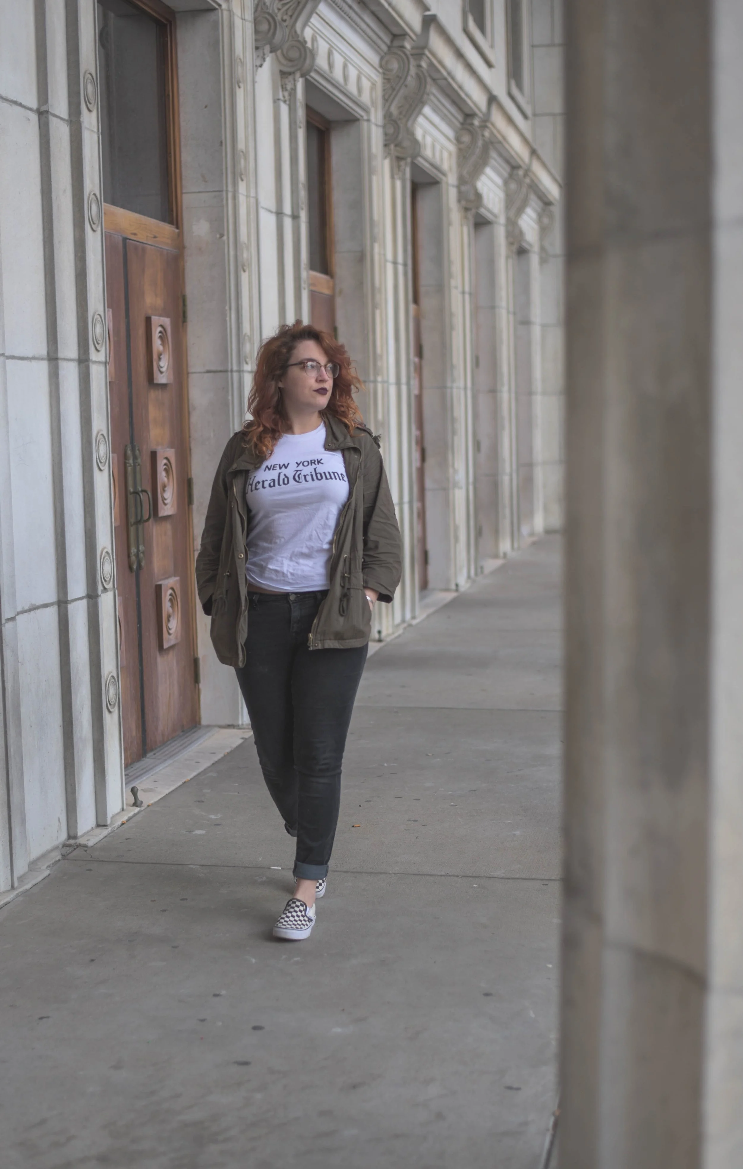 Red headed woman with curly hair, tortoiseshell glasses, and dark lipstick walking past a limestone building's facade. She's wearing a white New York Herald Tribune tee, a hunter green jacket, black jeans, and checkered slip-on Vans.