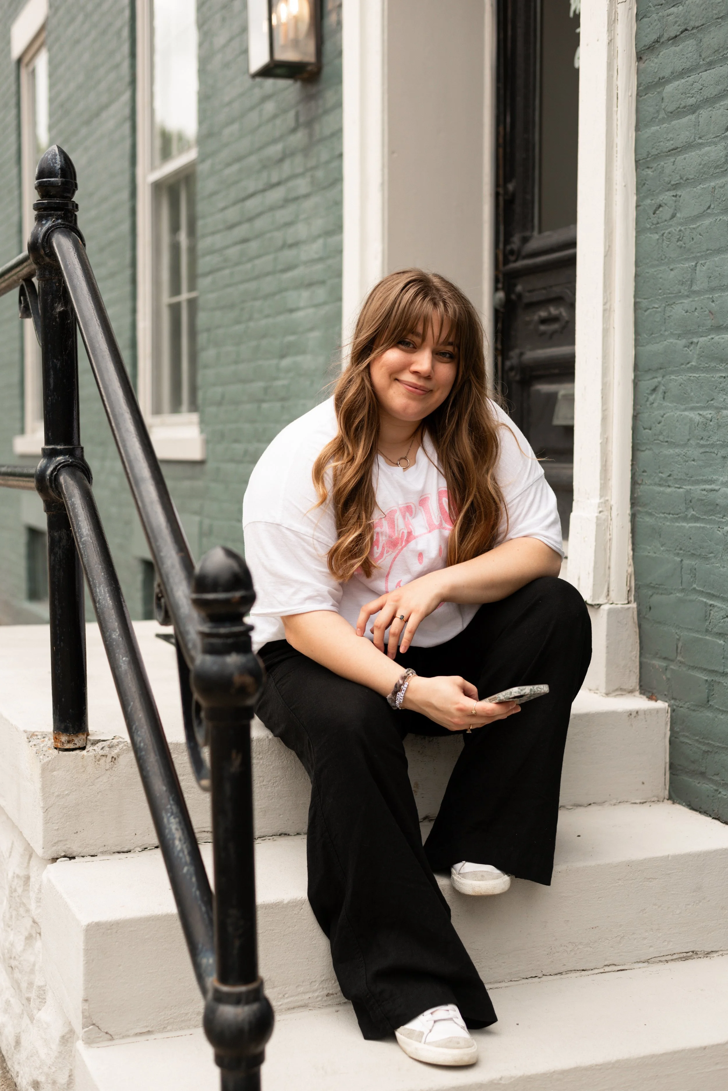a woman with long brown hair and bangs sits on steps outside a blue brick building in downtown evansville, indiana