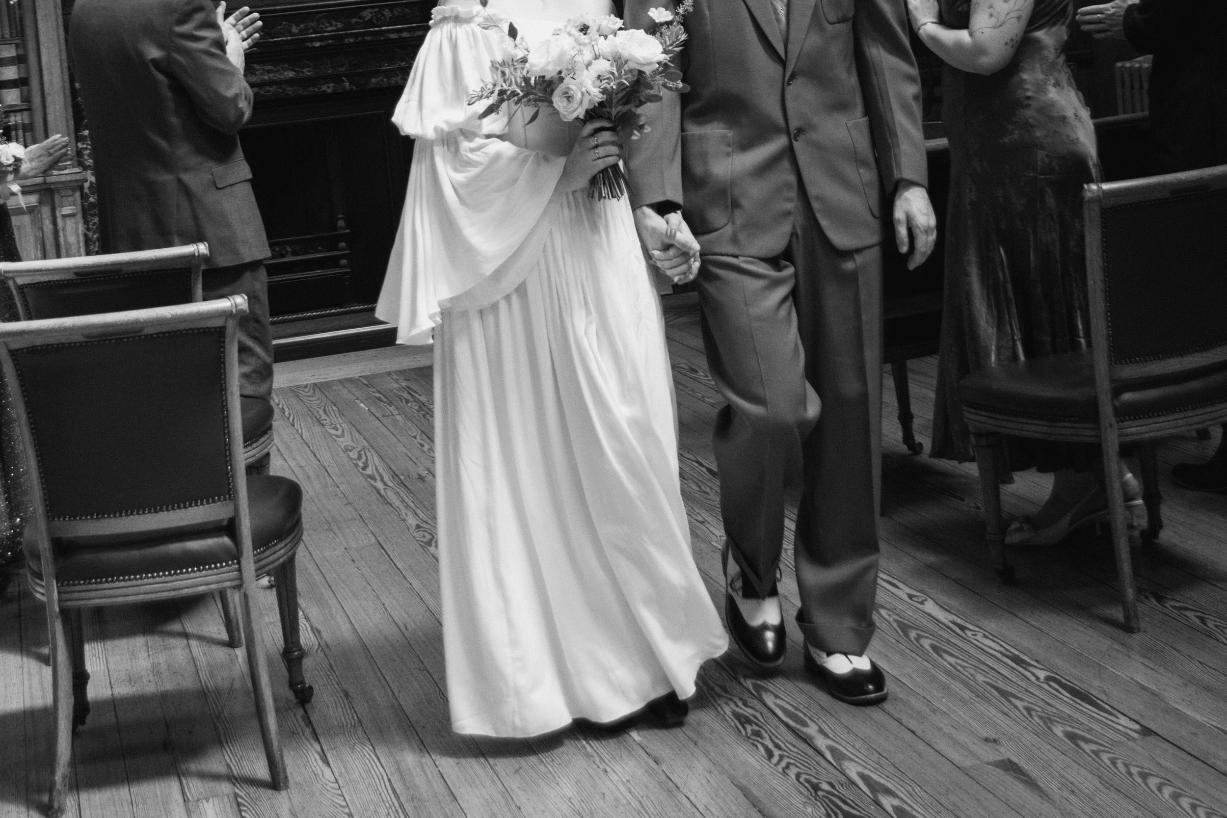 black and white image of a bride and groom walking back down the aisle after their "i do's". She is holding her bouque and they are holding hands as they exit.