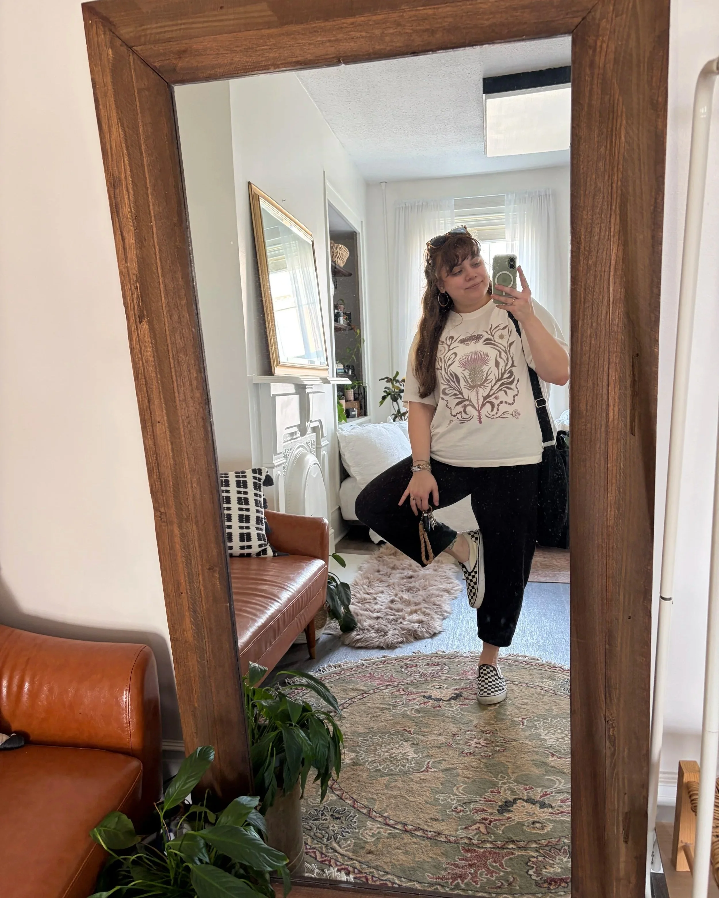 A brunette woman with bangs stands on one leg facing the mirror, taking a selfie with her phone. She's in a photo studio set up to feel like a bedroom, and you can see a warm-toned leather couch in the corner. She's holding her keys and smiling.
