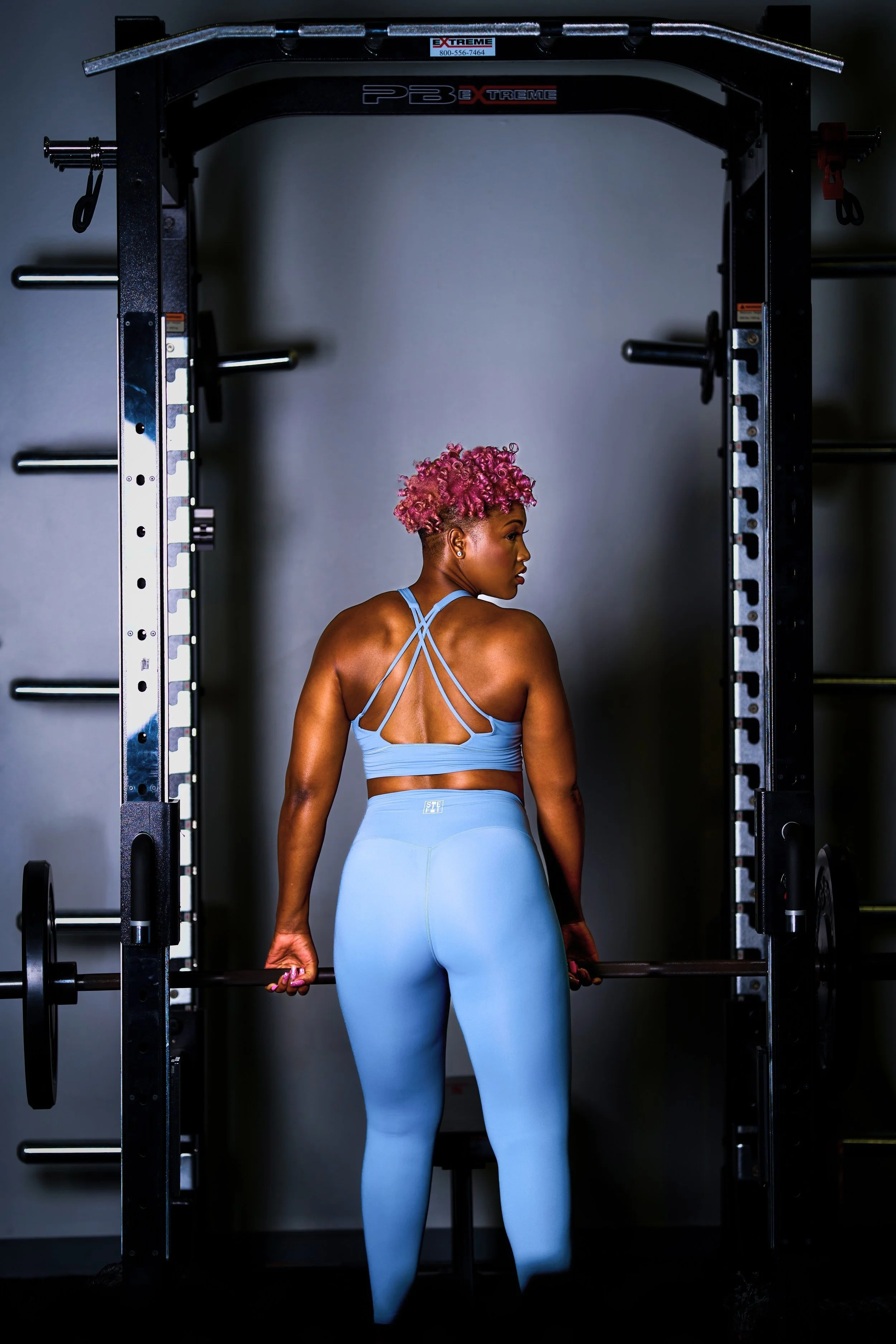 A woman with pink curly hair wearing light blue workout clothes holding a barbell in a gym, standing in front of a black power rack.