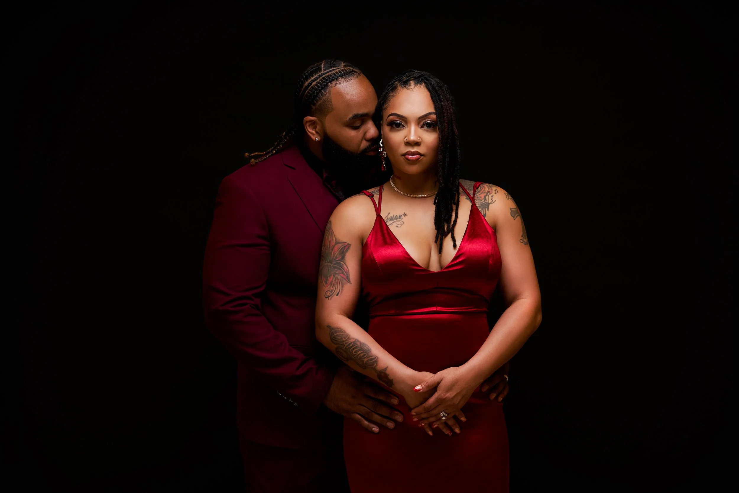 A man and woman dressed in matching burgundy outfits pose closely against a black background. The man, with braided hair and a beard, stands behind the woman, who has long, dreadlocked hair and tattoos on her arms. They are holding hands in front of
