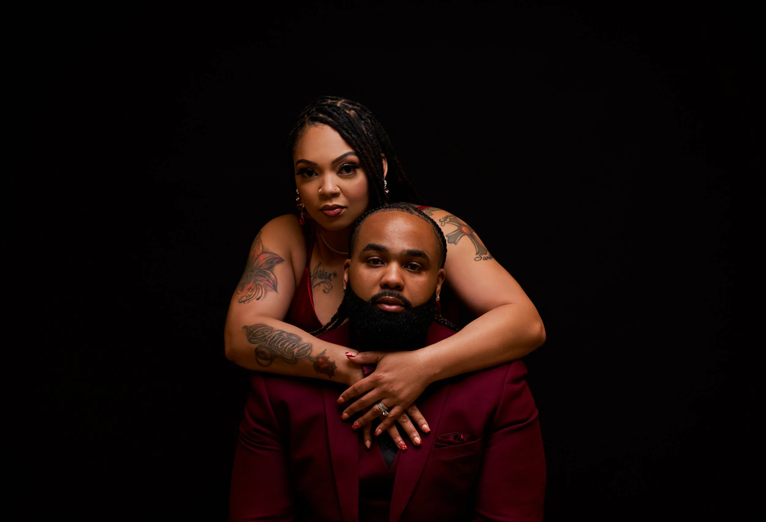 A woman with tattoos and makeup embraces a man with a beard, both dressed in burgundy outfits, against a black background.