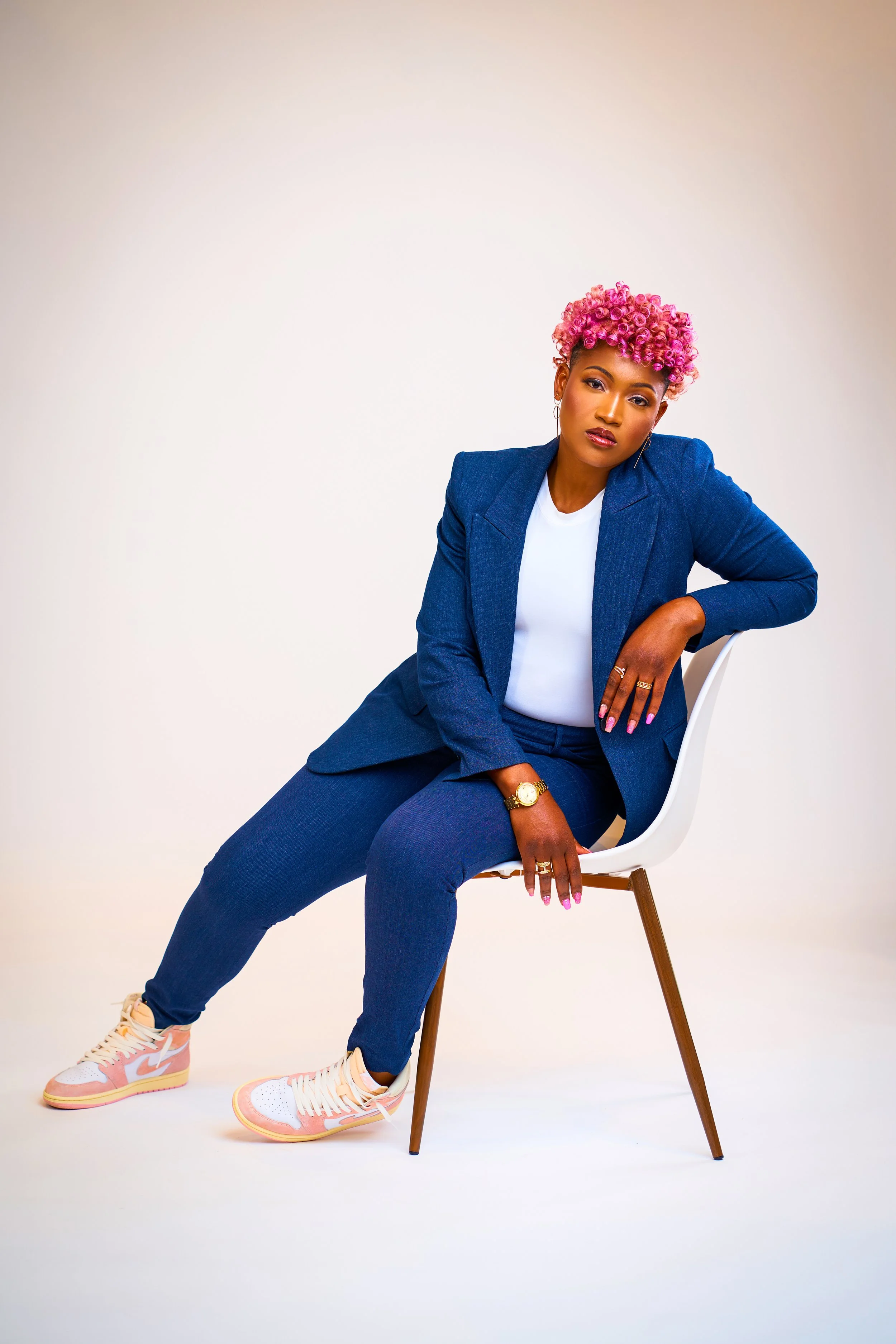 A woman with pink curly hair wearing a blue suit and white t-shirt, seated on a white chair with wooden legs, against a plain light background.