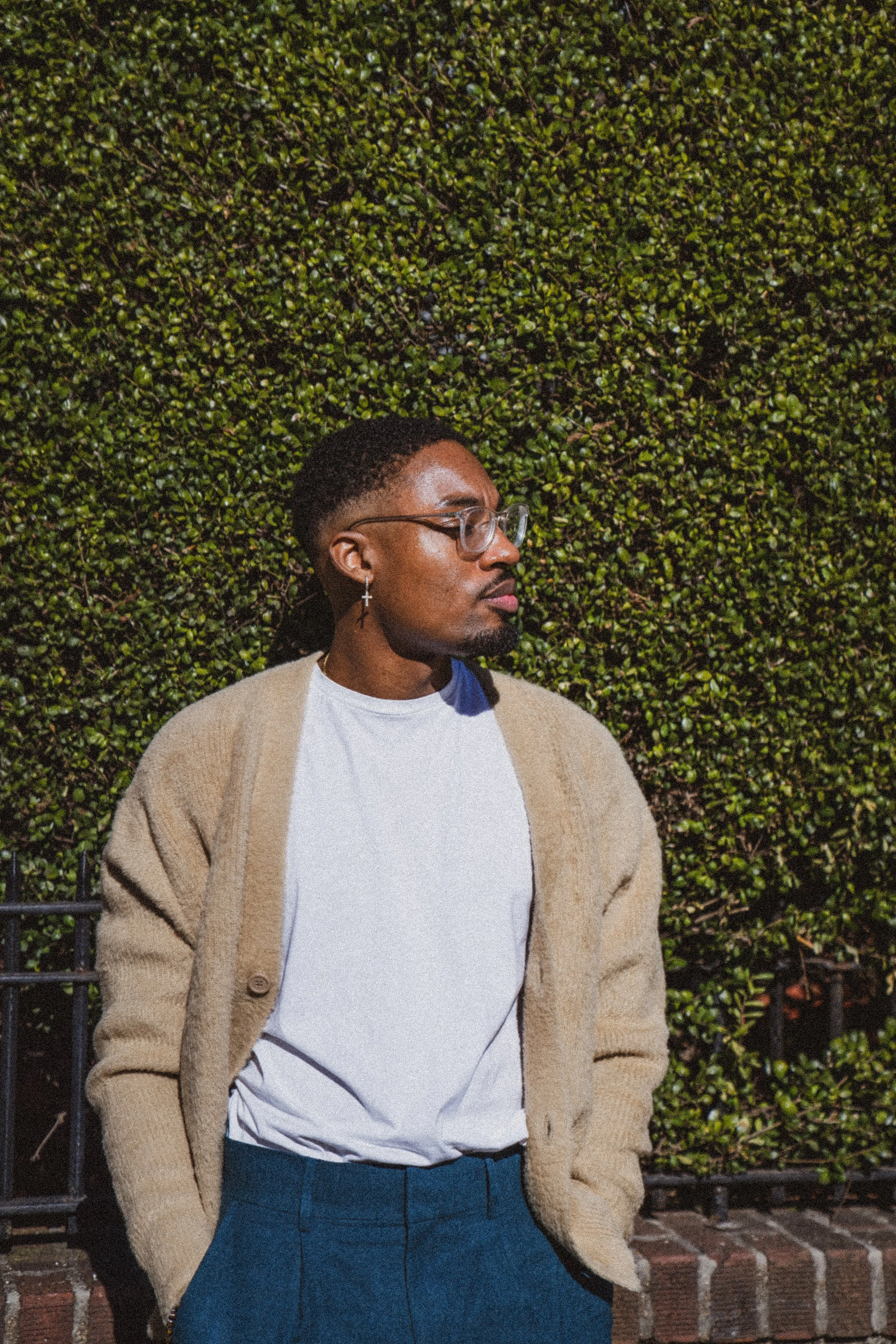 A man standing outdoors in front of a green hedge, wearing glasses, a cross earring, a white shirt, a beige cardigan, and blue pants.