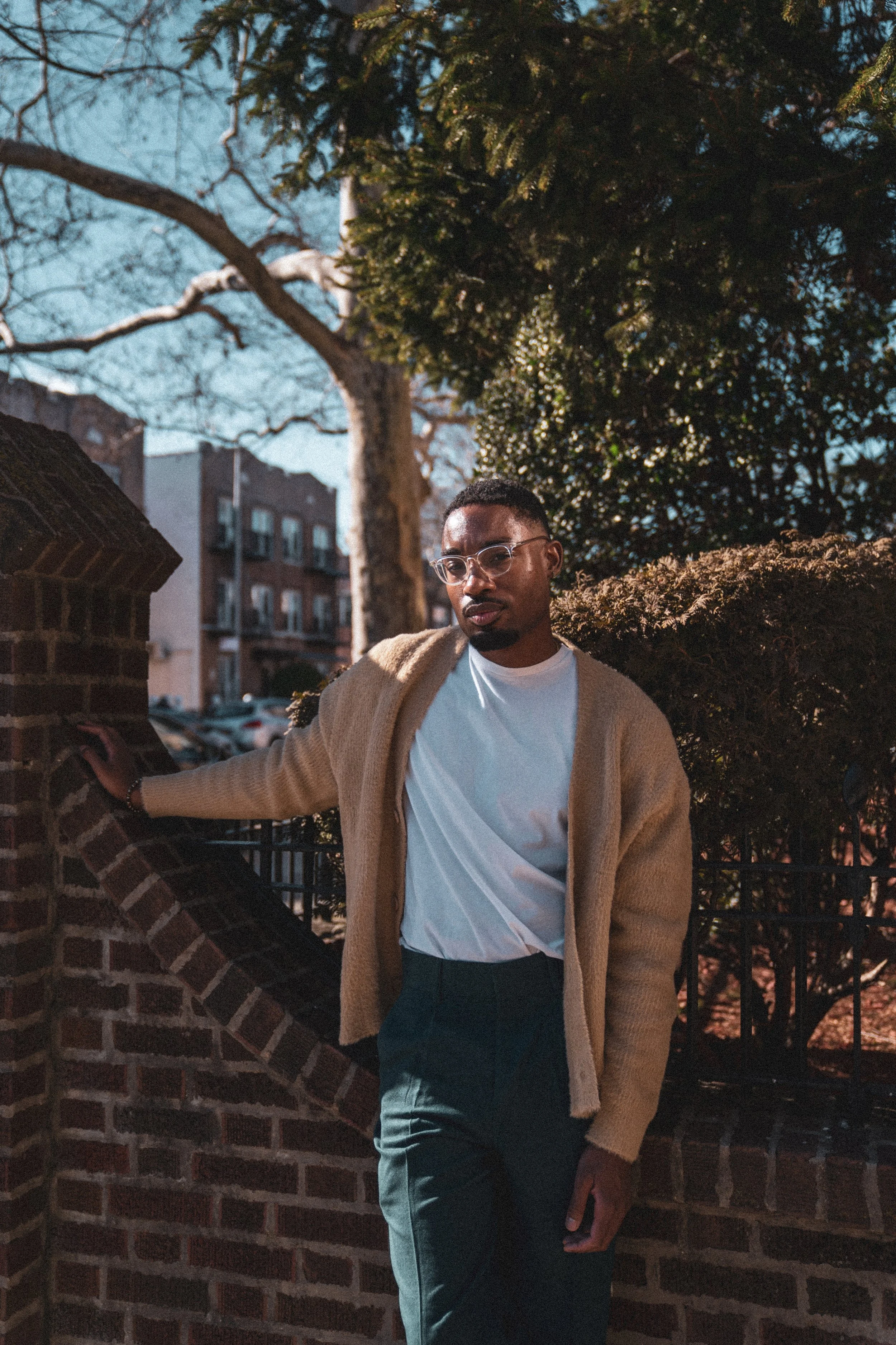 A young man with glasses wearing a white t-shirt and tan cardigan, leaning against a brick wall outdoors with trees and buildings in the background during daytime.