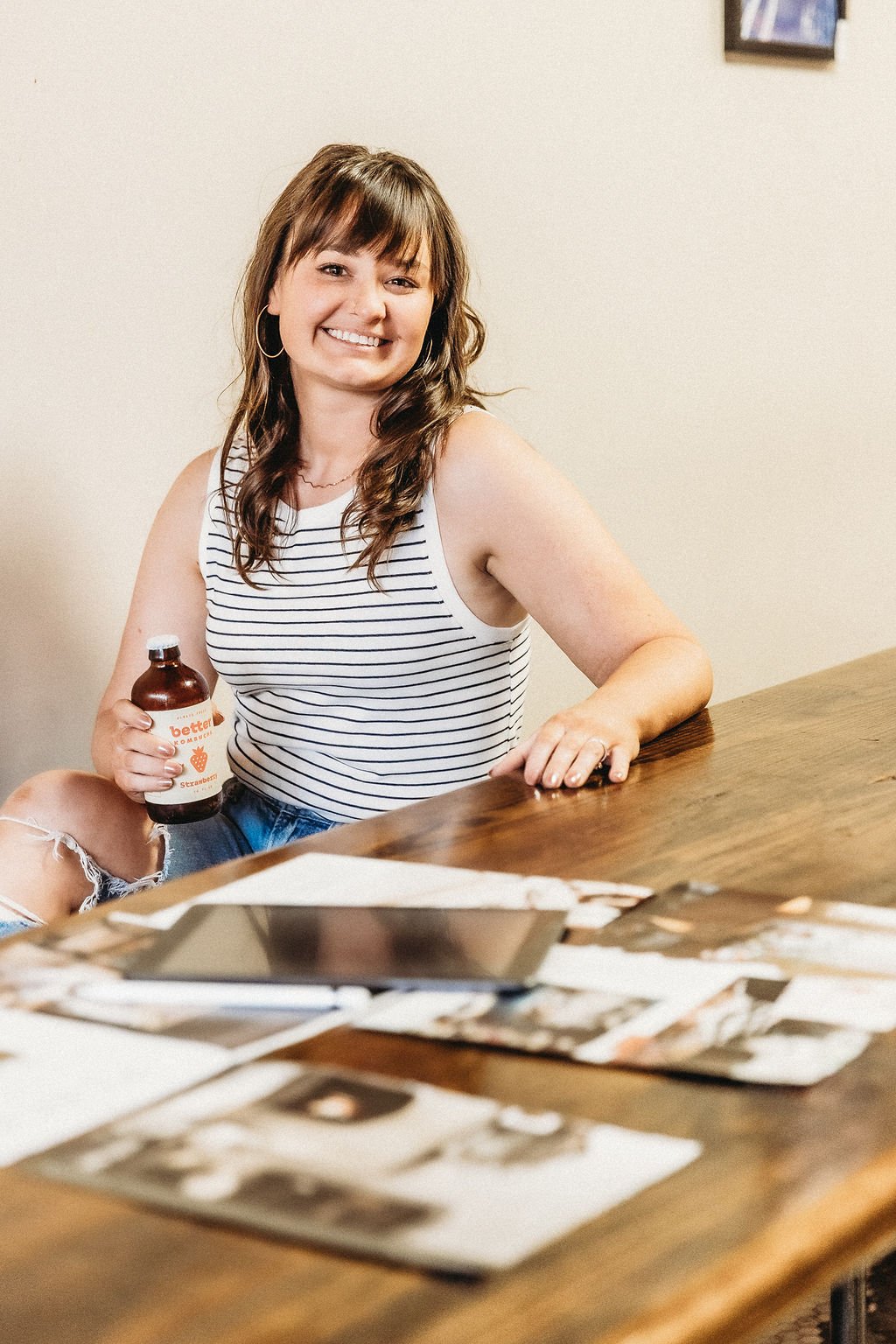 A woman with shoulder-length brown hair, wearing a white and navy striped sleeveless top and ripped denim shorts, smiling and holding a bottle of kombucha, sitting at a wooden table with photographs and a tablet in front of her.