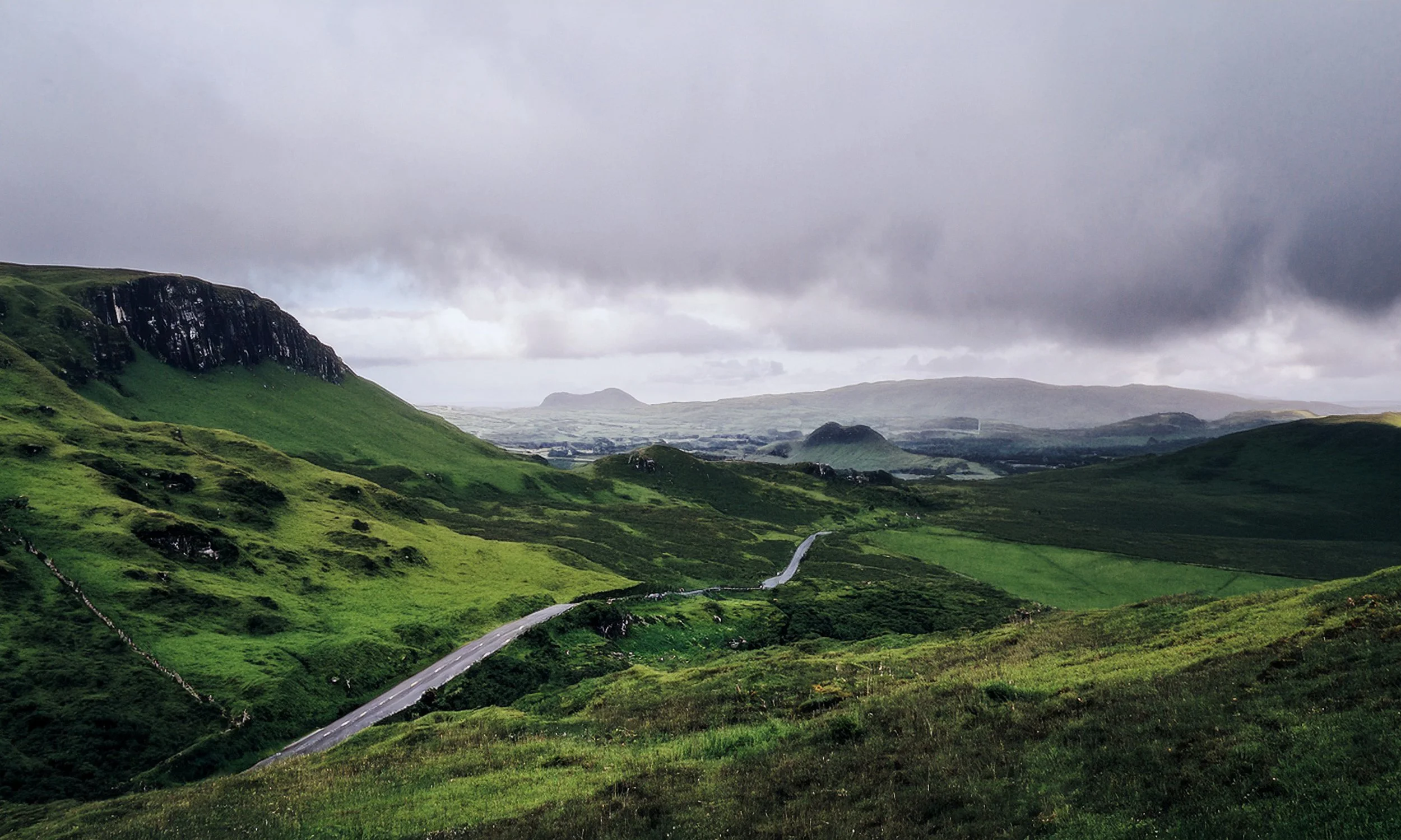 Green rolling hills with a winding road and a cloudy sky over a rural landscape.