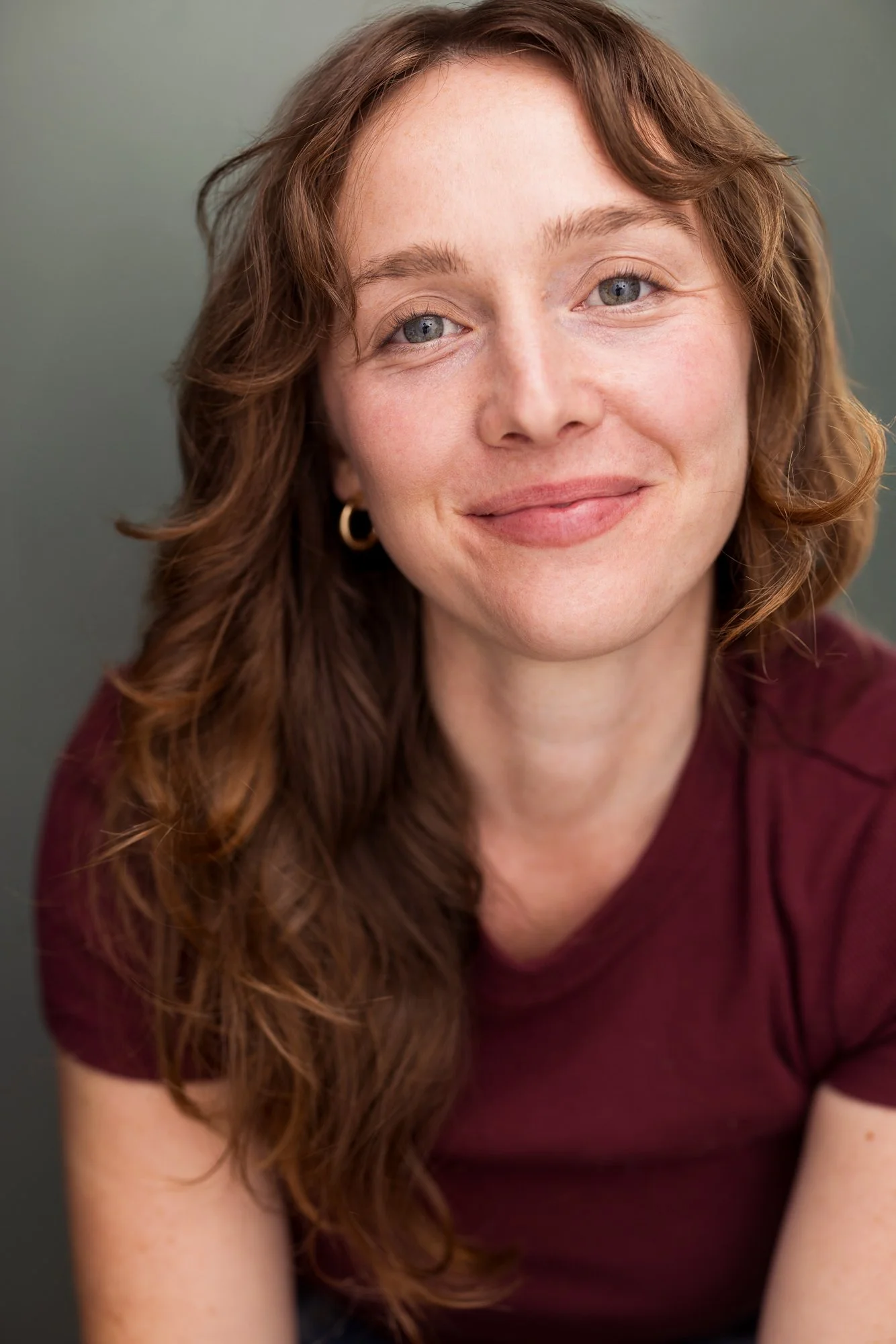 A red haired actress with green eyes, Emma K. Miles, smiles while wearing a maroon shirt and small hoop earrings, against a neutral background.