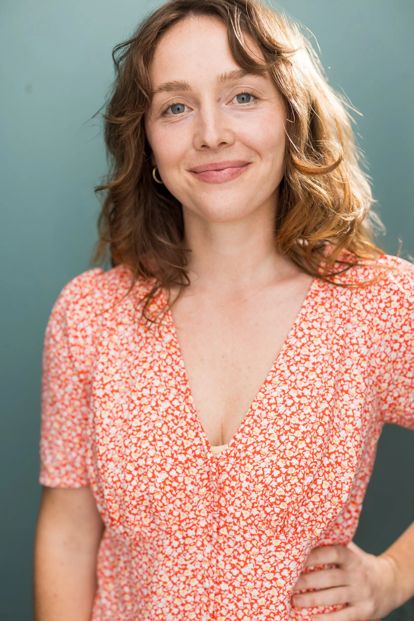 Actress Emma K. Miles with wavy, shoulder-length red hair, green eyes, and fair freckled skin, smiling and wearing a coral and white patterned dress.