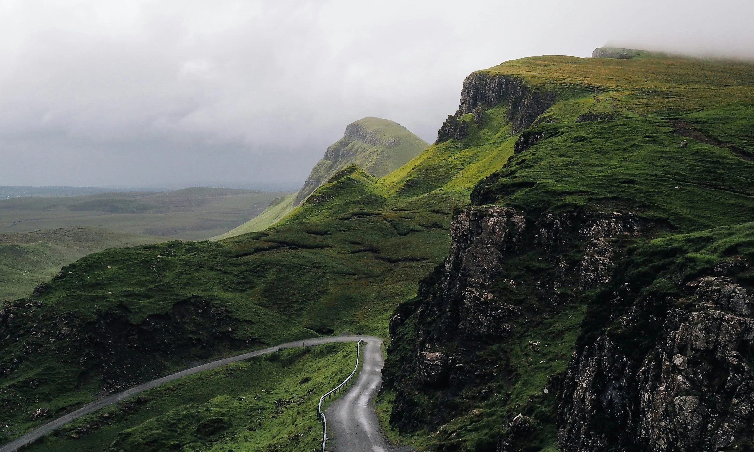 A winding road running through green, rugged hills with steep cliffs and low clouds in the background.