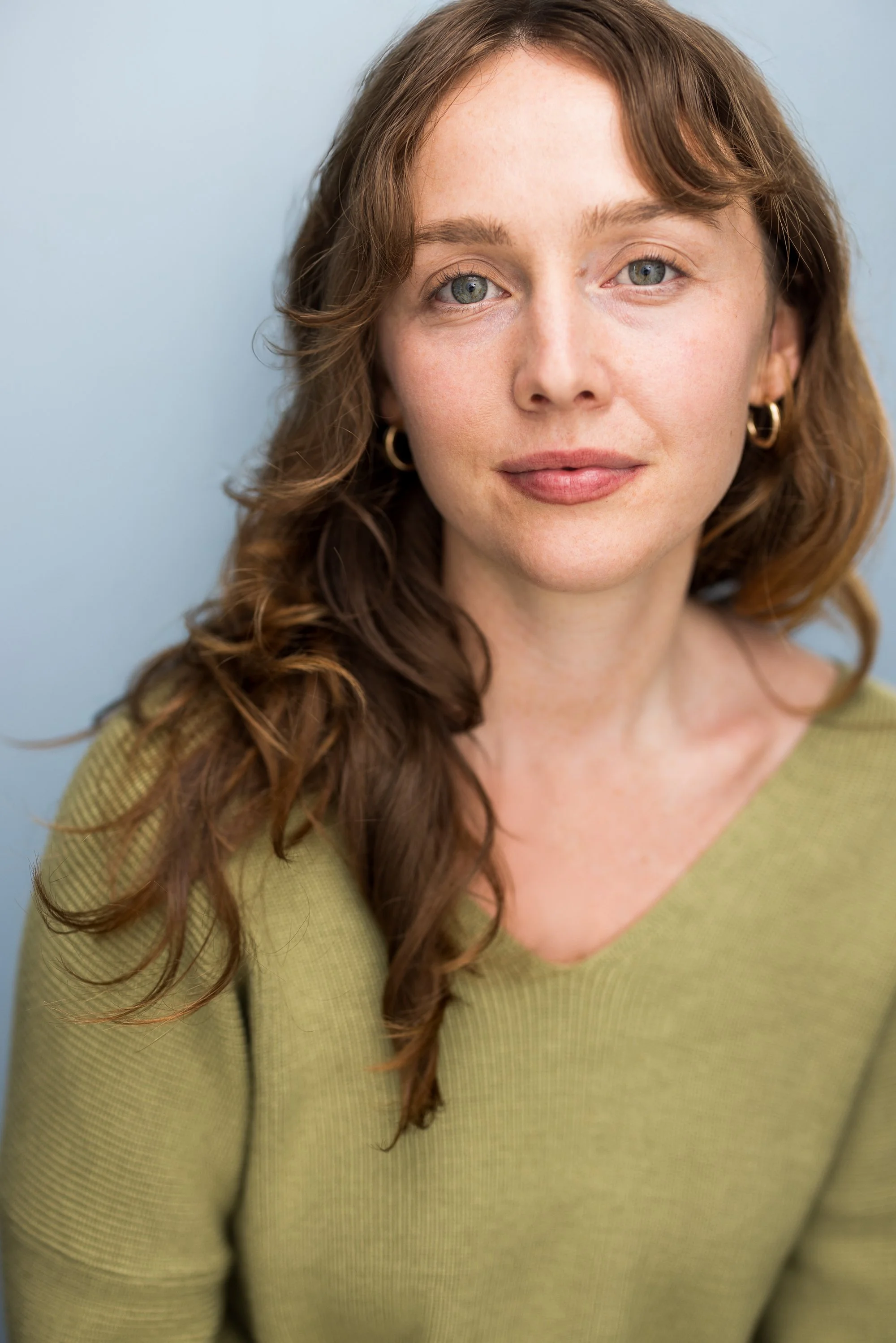 Close-up portrait of Emma K. Miles, with green eyes and wavy auburn hair, wearing gold hoop earrings and a light green top, against a light blue background.
