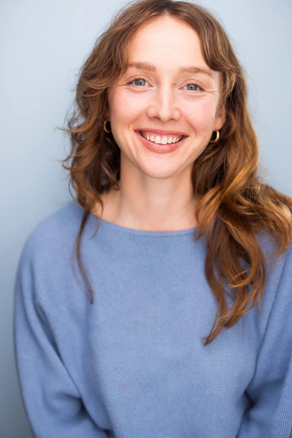Portrait of Emma K. Miles, a woman with long red auburn hair, green eyes, wearing a blue sweater, smiling against a light blue background.