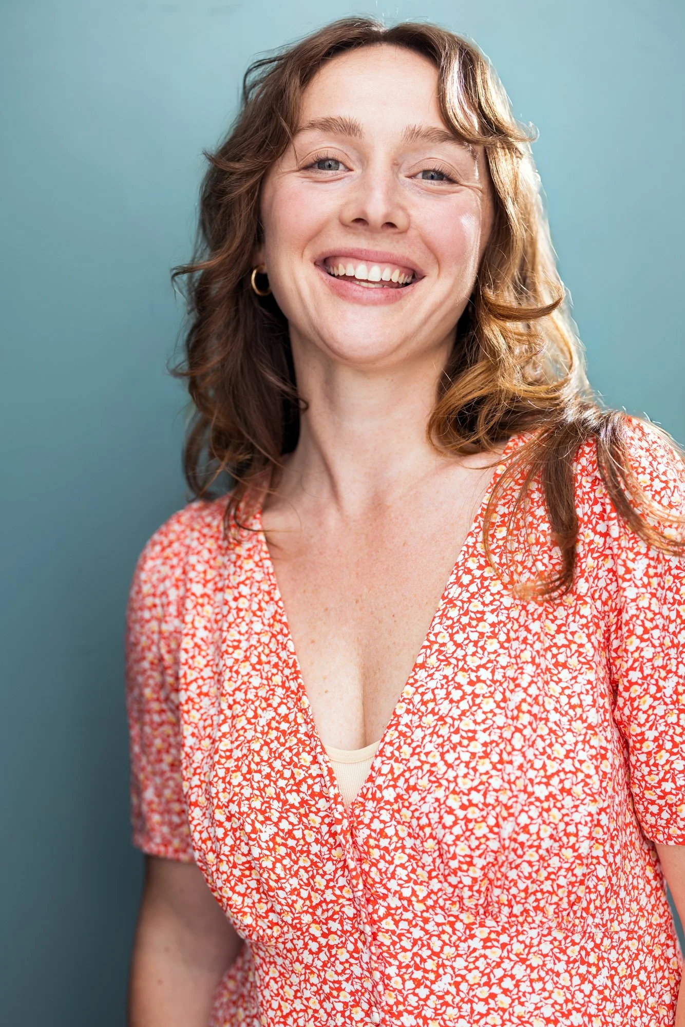 Emma K. Miles, a woman actress,  with fair skin and curly, shoulder-length auburn hair, smiling, wearing a peach and white floral patterned top and gold hoop earrings, standing against a light blue background.