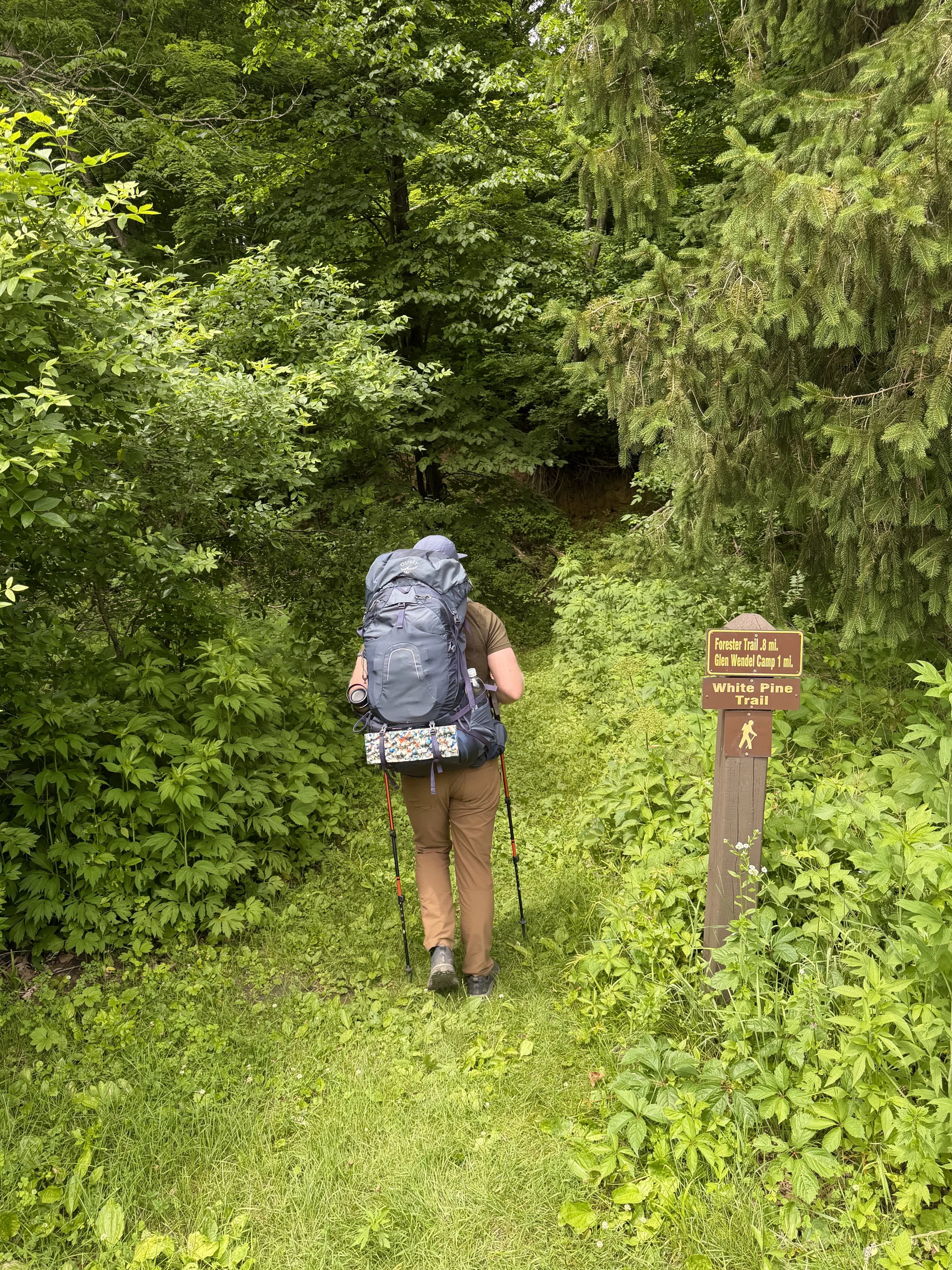 A hiker with a large backpack and brown beanie walking on a dirt trail in a wooded area during fall, with a small wooden bridge in the background.