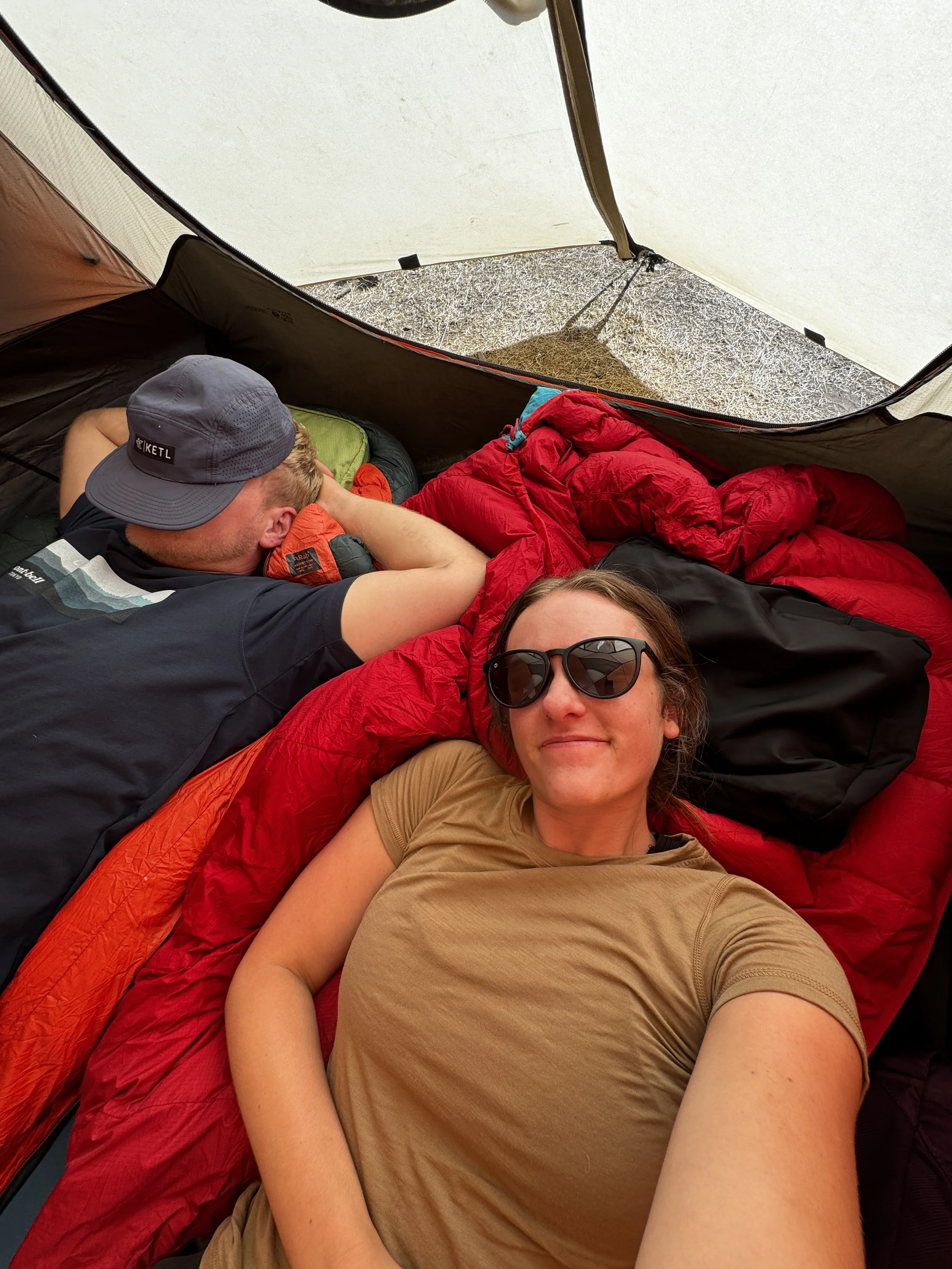 A person wearing a Patagonia hat, a black jacket, and a black shirt is resting their head on a green and yellow sleeping bag inside a tent. The background shows a forest with leafless trees.