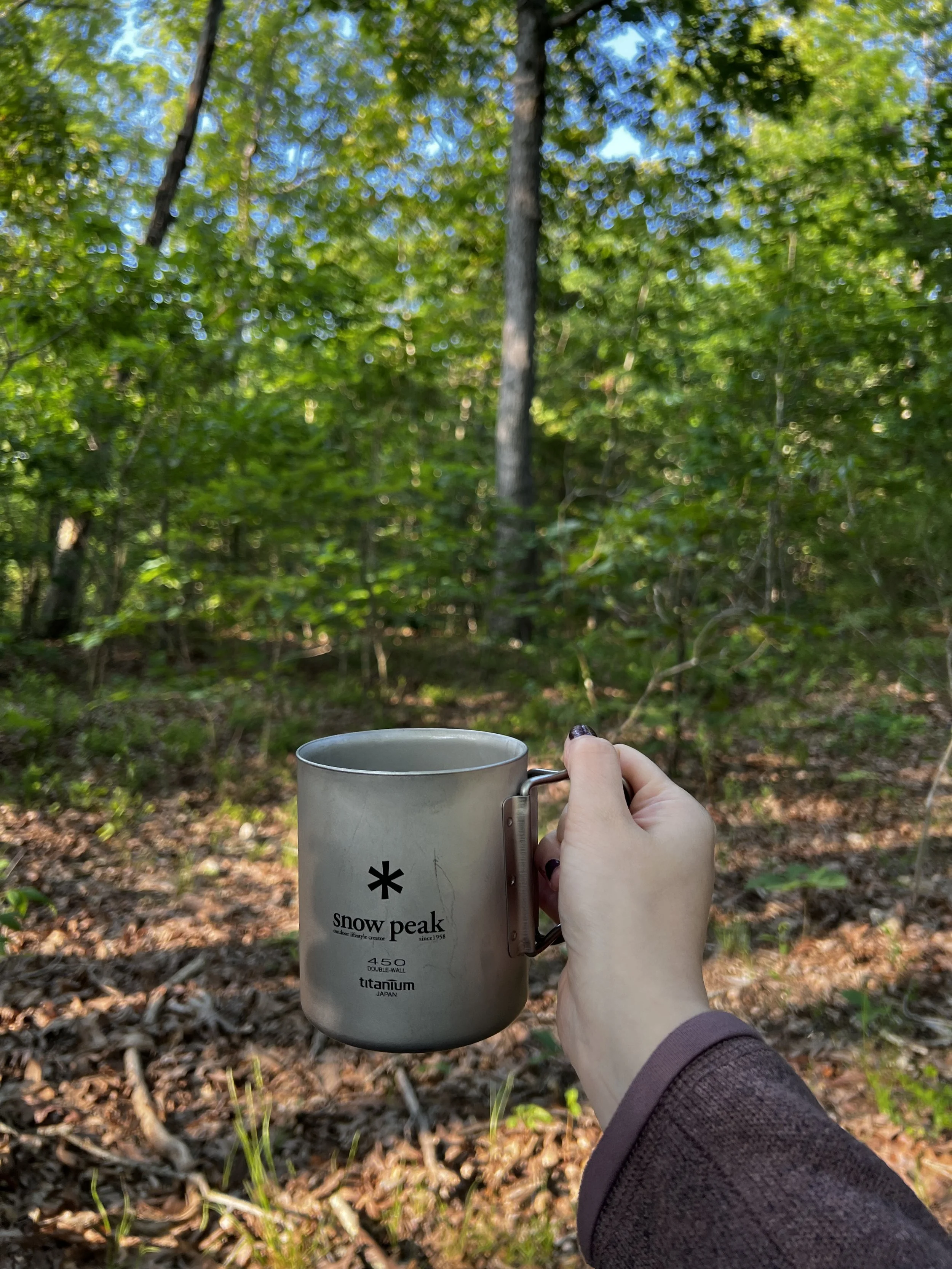 A man wearing a yellow and green Patagonia cap and a black shirt that says 'Leprechaun Chase' is drinking from a gray mug in a wooded area.