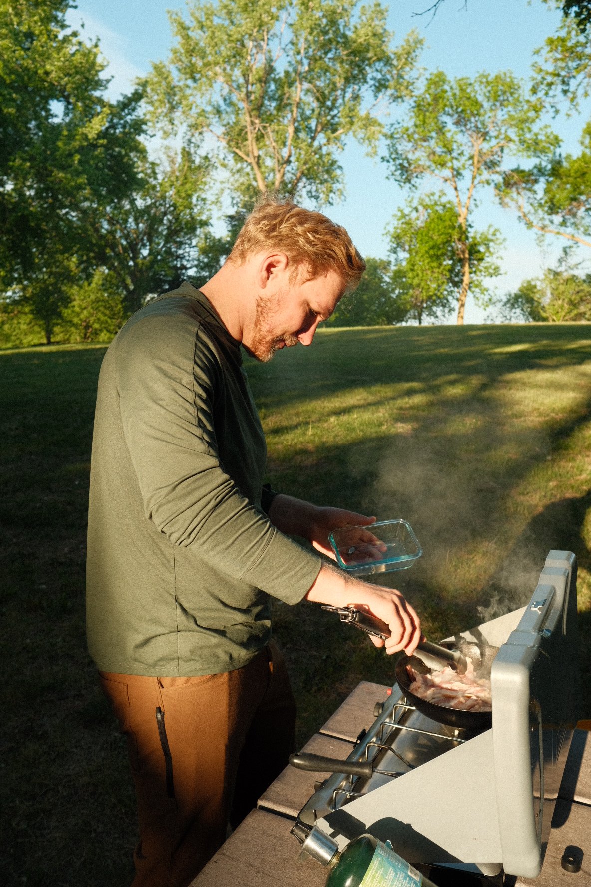 A man sitting in a camp chair outdoors, cooking breakfast on a portable stove, with a coffee pot and frying eggs. There are trees and a clear sky in the background, and a hand holding a coffee mug in the foreground. Text overlay reads, 'Prepping for the day with coffee and eggs' with a location tag, 'Grand Canyon South Rim'.
