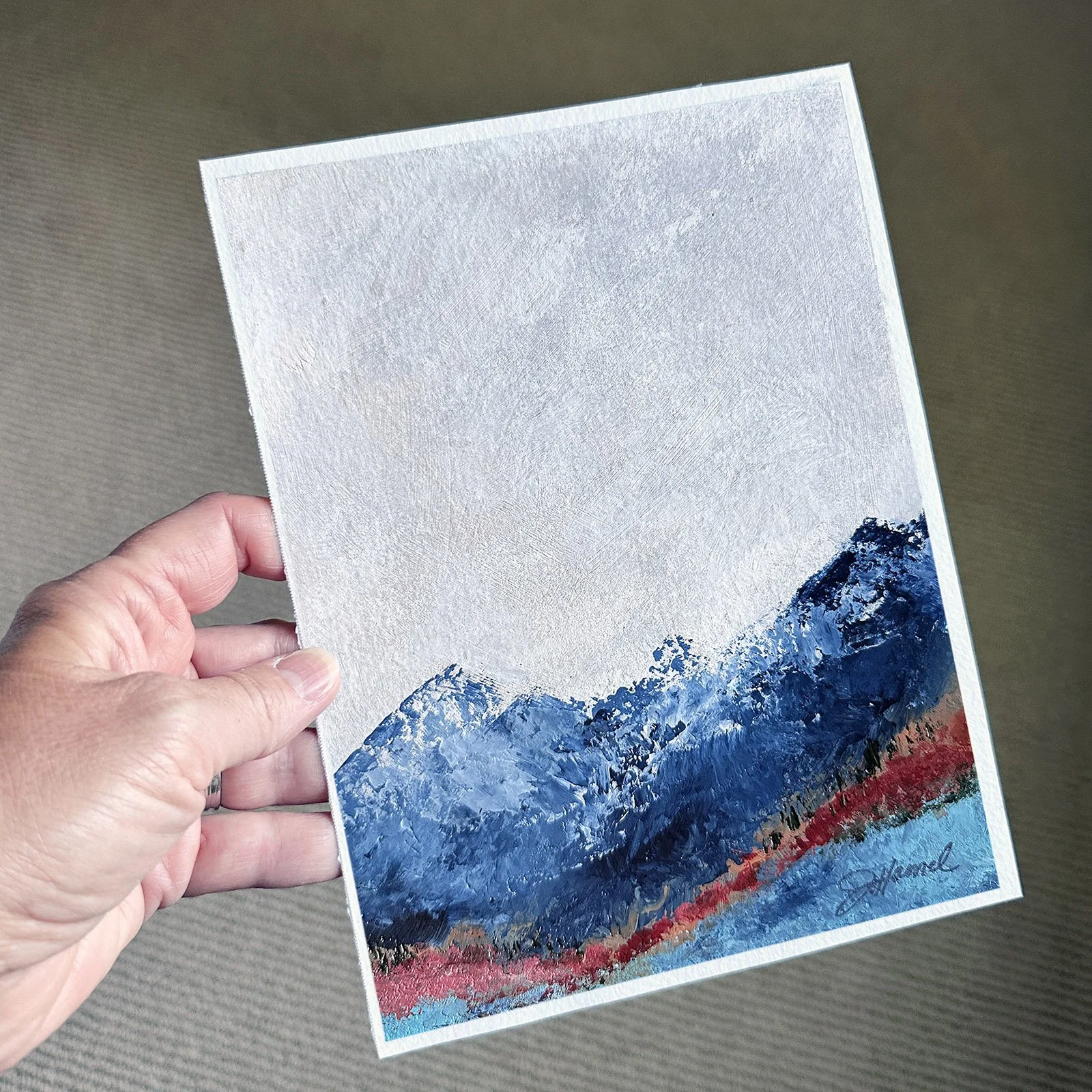 Hand holding a small blue mountain landscape painting.