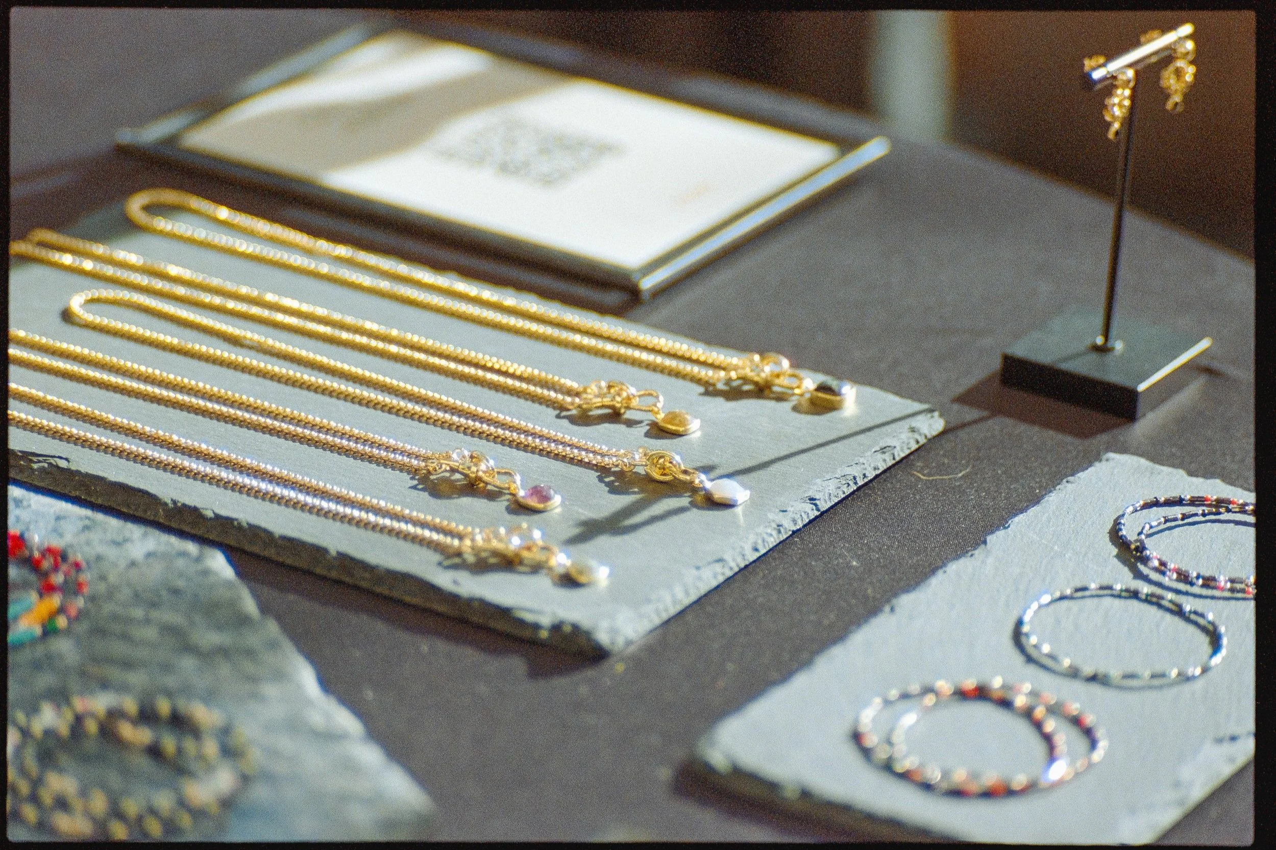 Various gold and silver jewelry including necklaces, bracelets, and earrings displayed on a black and grey table, with additional jewelry pieces in the background.