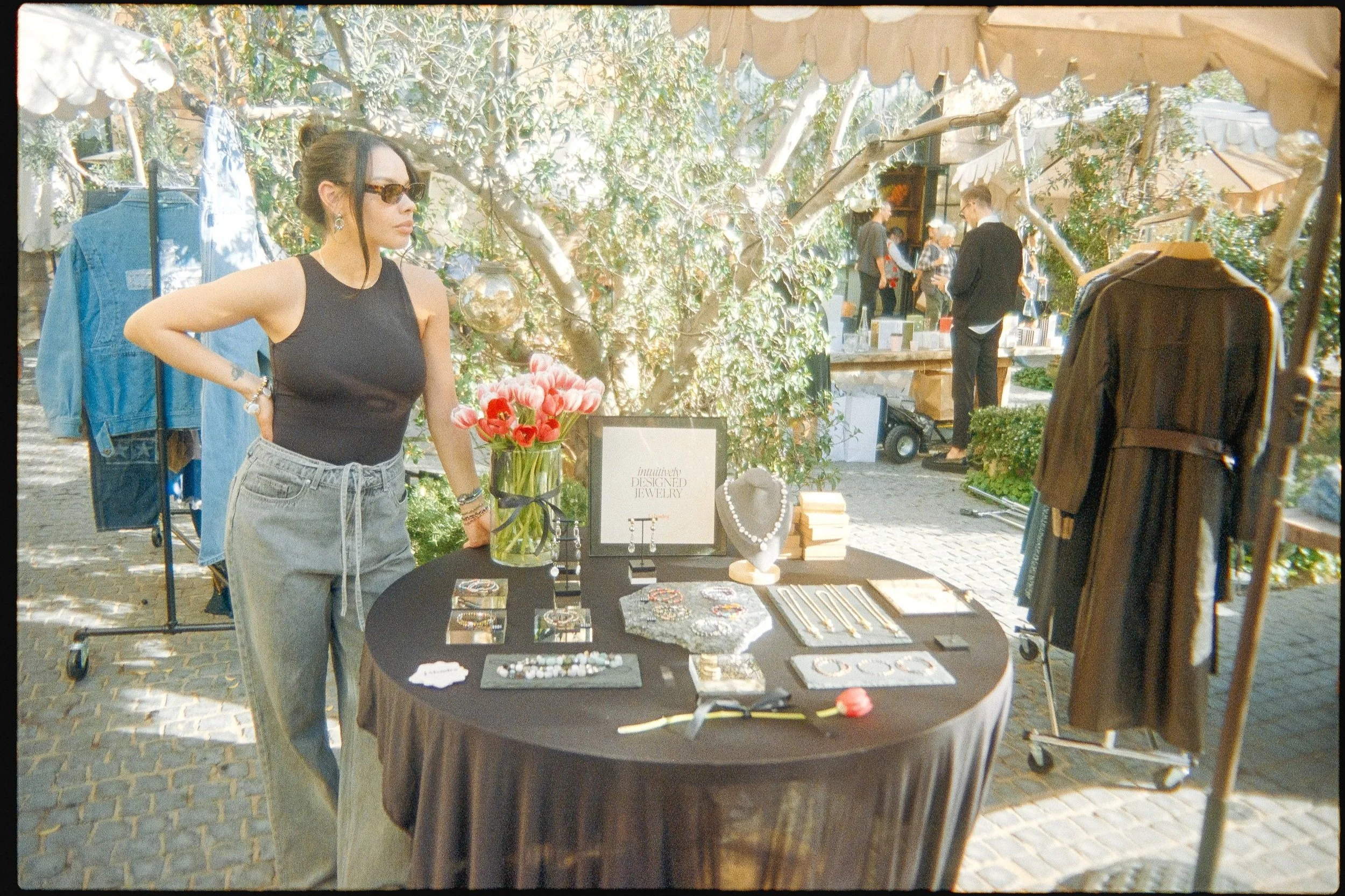 A woman with dark hair and sunglasses, wearing a black sleeveless top and gray pants, stands at an outdoor jewelry stall with a black tablecloth, displaying jewelry items like necklaces, earrings, and bracelets. The stall has a vase of pink tulips, a