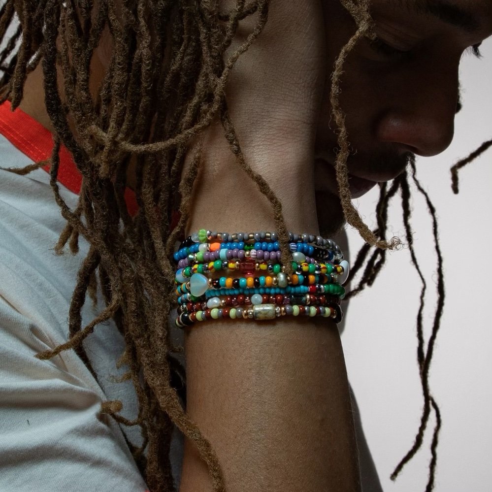 Close-up of a person with long dreadlocks, wearing multiple colorful beaded bracelets on their wrist, resting their chin on their arm, with a neutral background.