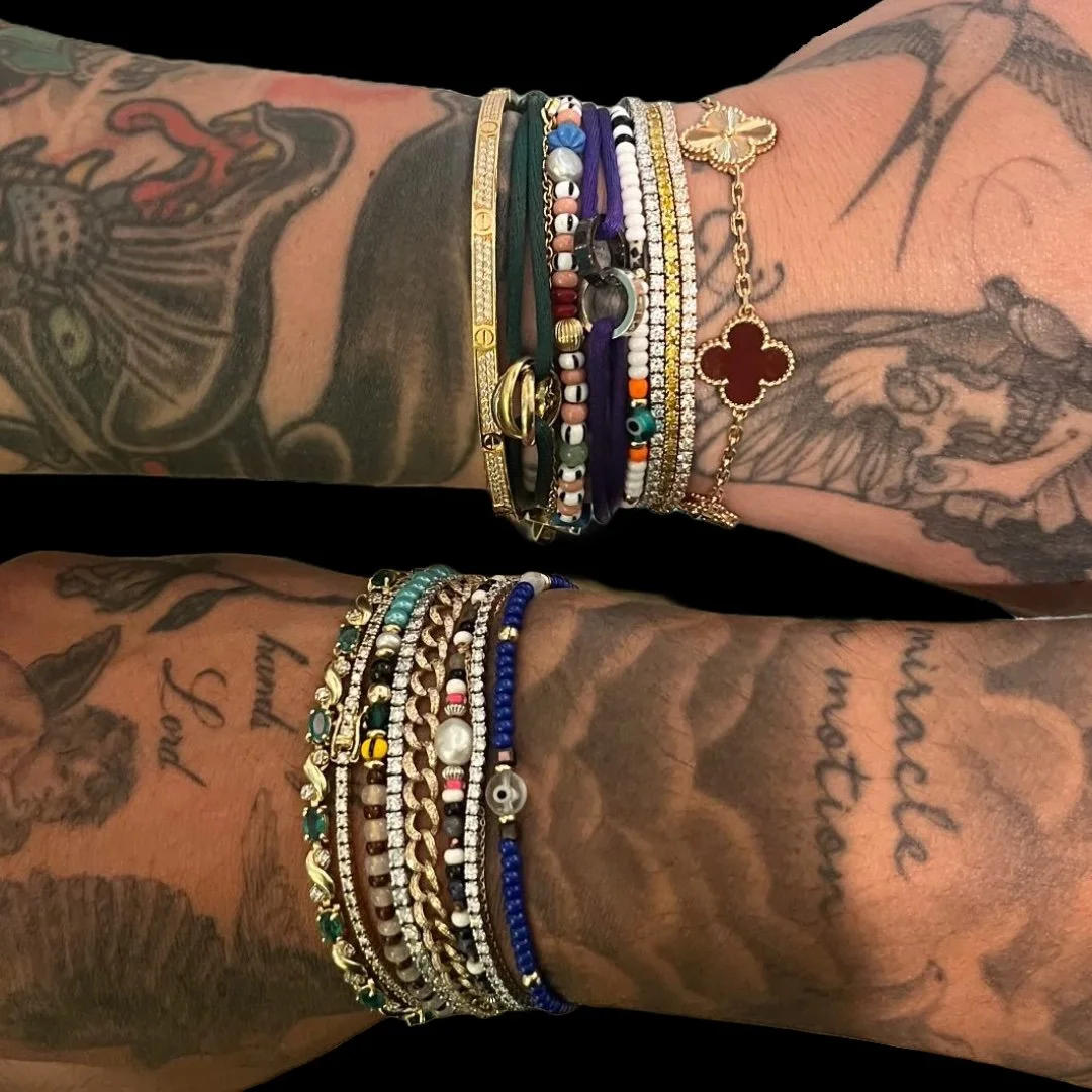Close-up of two tattooed arms adorned with multiple silver, gold, beaded, and charm bracelets, showcasing diverse styles.