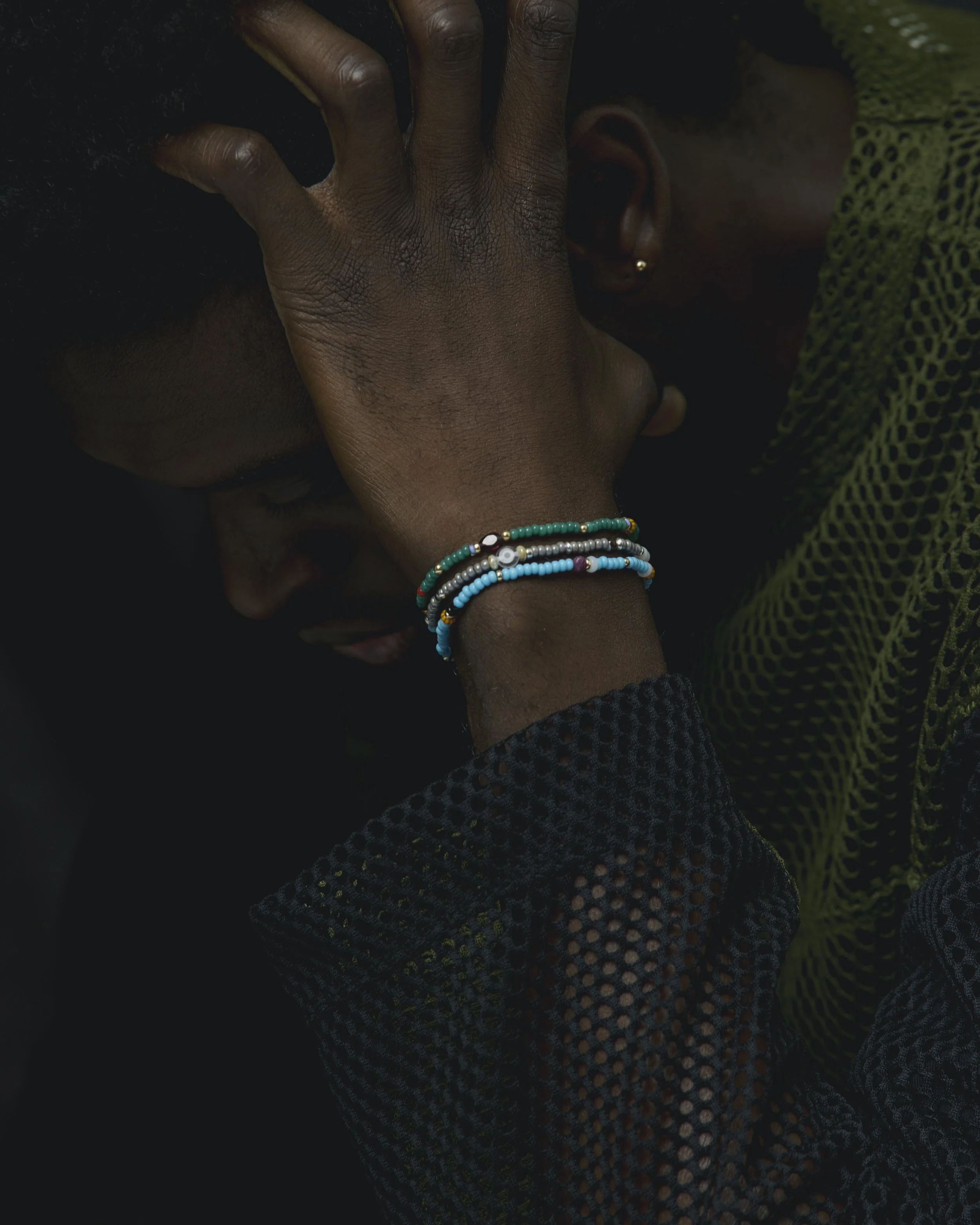 Two people with dark skin, one with short hair and one with a headscarf, sharing an intimate moment with their foreheads touching and eyes closed. The person in the foreground is wearing several colorful beaded bracelets.