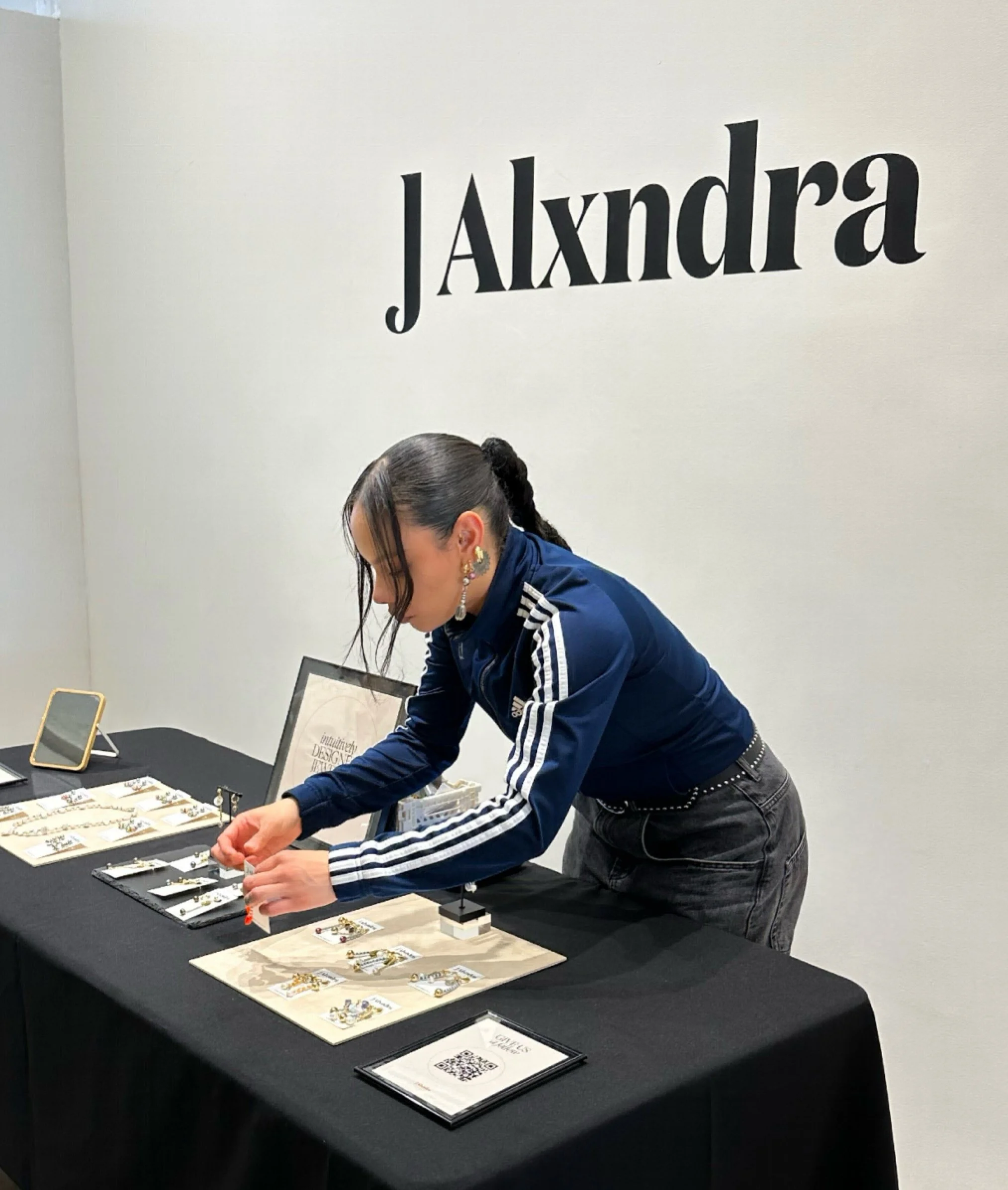A woman with dark hair in a ponytail, wearing a blue Adidas jacket with white stripes and earrings, is arranging jewelry on a black table at a booth with a sign that says 'JAlxndra.'