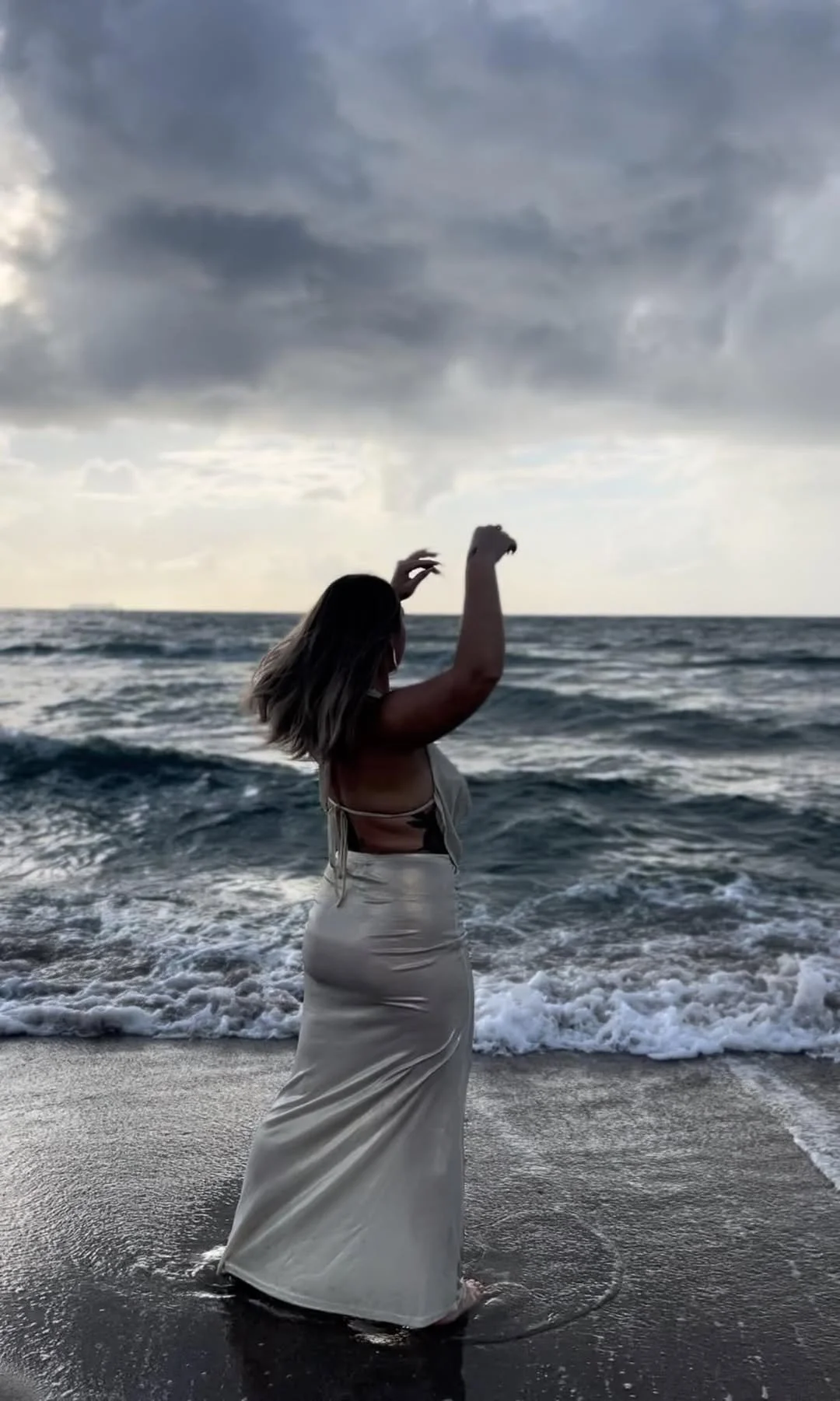 Woman in a flowing dress wading in the ocean, with dark, cloudy sky overhead