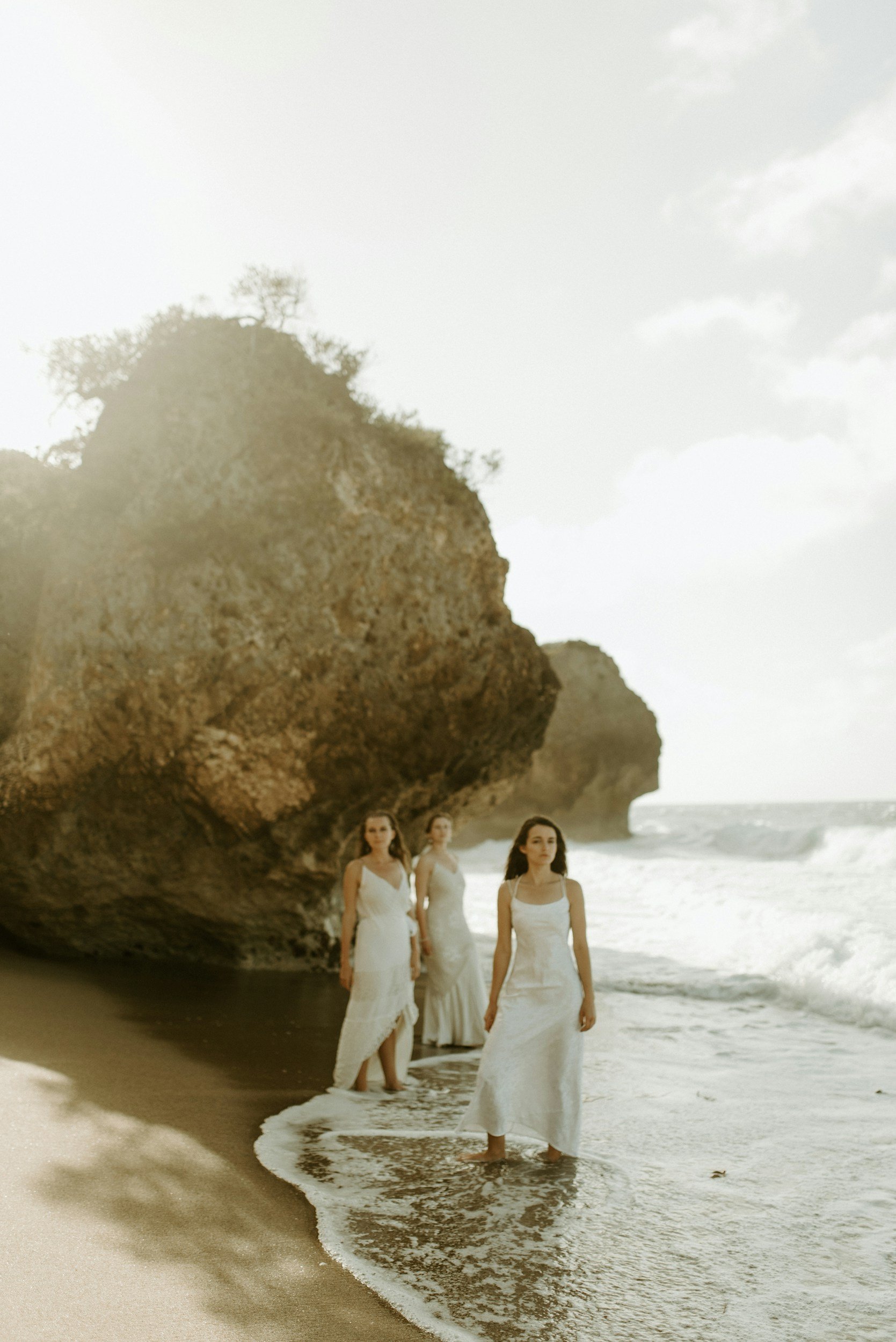 Three women in white dresses walking along the beach near large rock formations near the shoreline during daytime.
