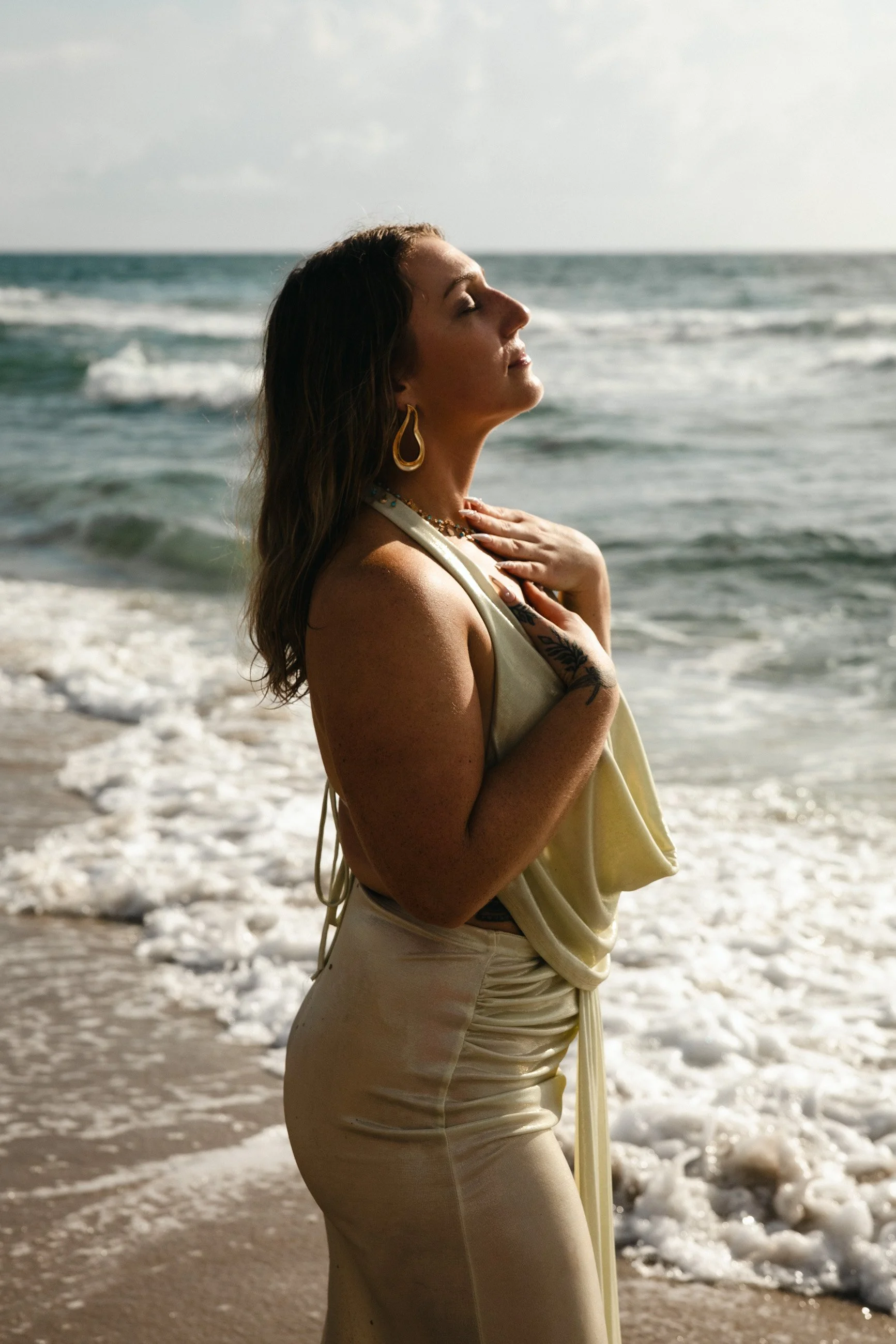 A woman standing on the beach near the ocean, with her eyes closed and one hand on her chest, wearing cream-colored clothing and gold jewelry.