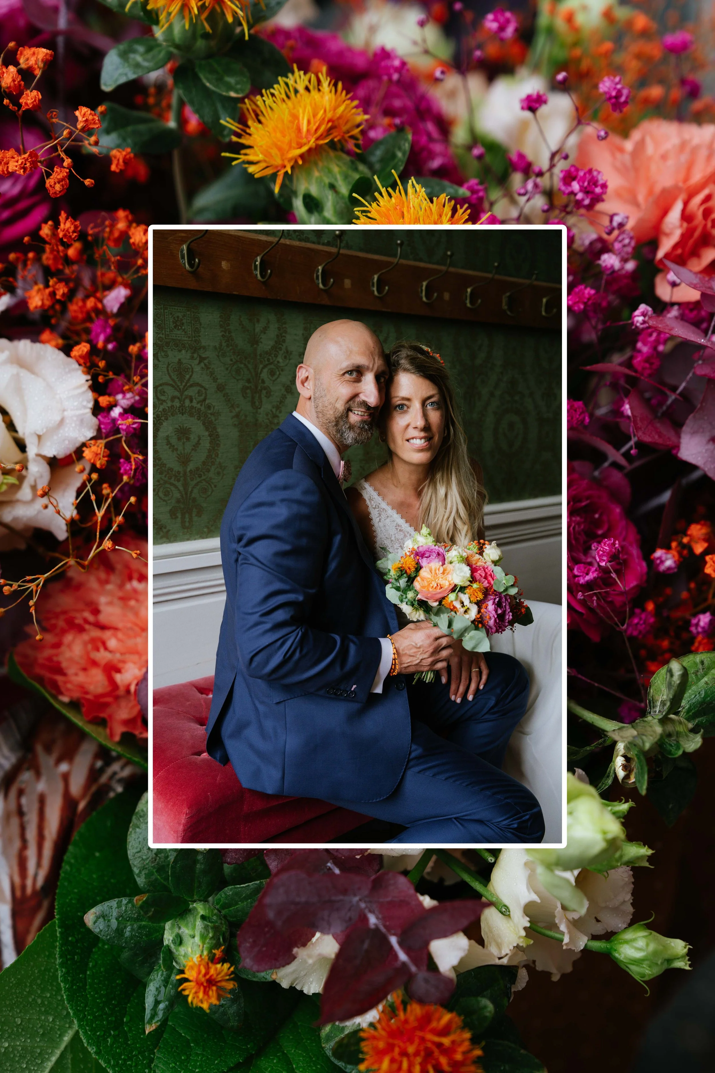 Une photo de mariage montrant un homme en costume bleu sombre et une femme avec une robe blanche, tenant un bouquet de fleurs colorées, assis dans une pièce avec un tapis rouge et un mur vert à motifs.