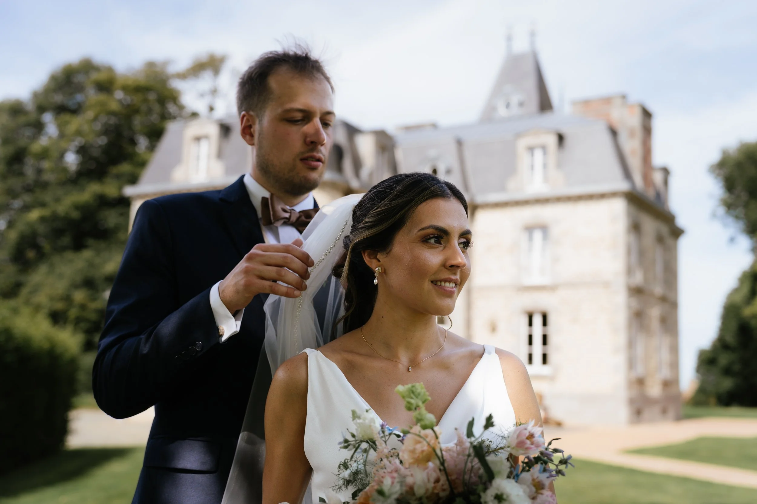 Un homme en costume ajustant le voile d'une femme en robe de mariage, tenant un bouquet de fleurs, devant un château en arrière-plan.