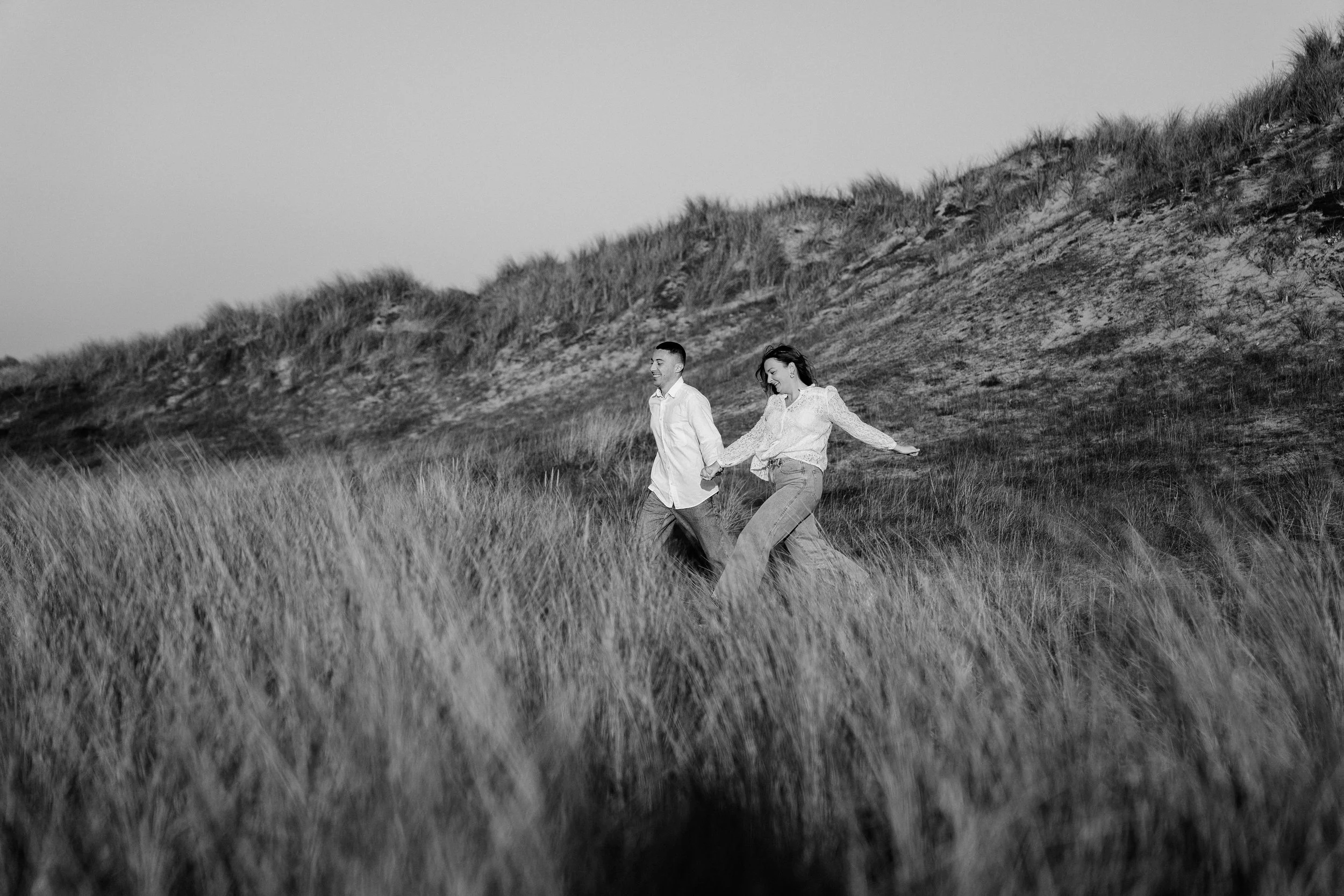 Un couple marche dans l'herbe haute sur un paysage de collines avec un ciel clair. L'image est en noir et blanc.