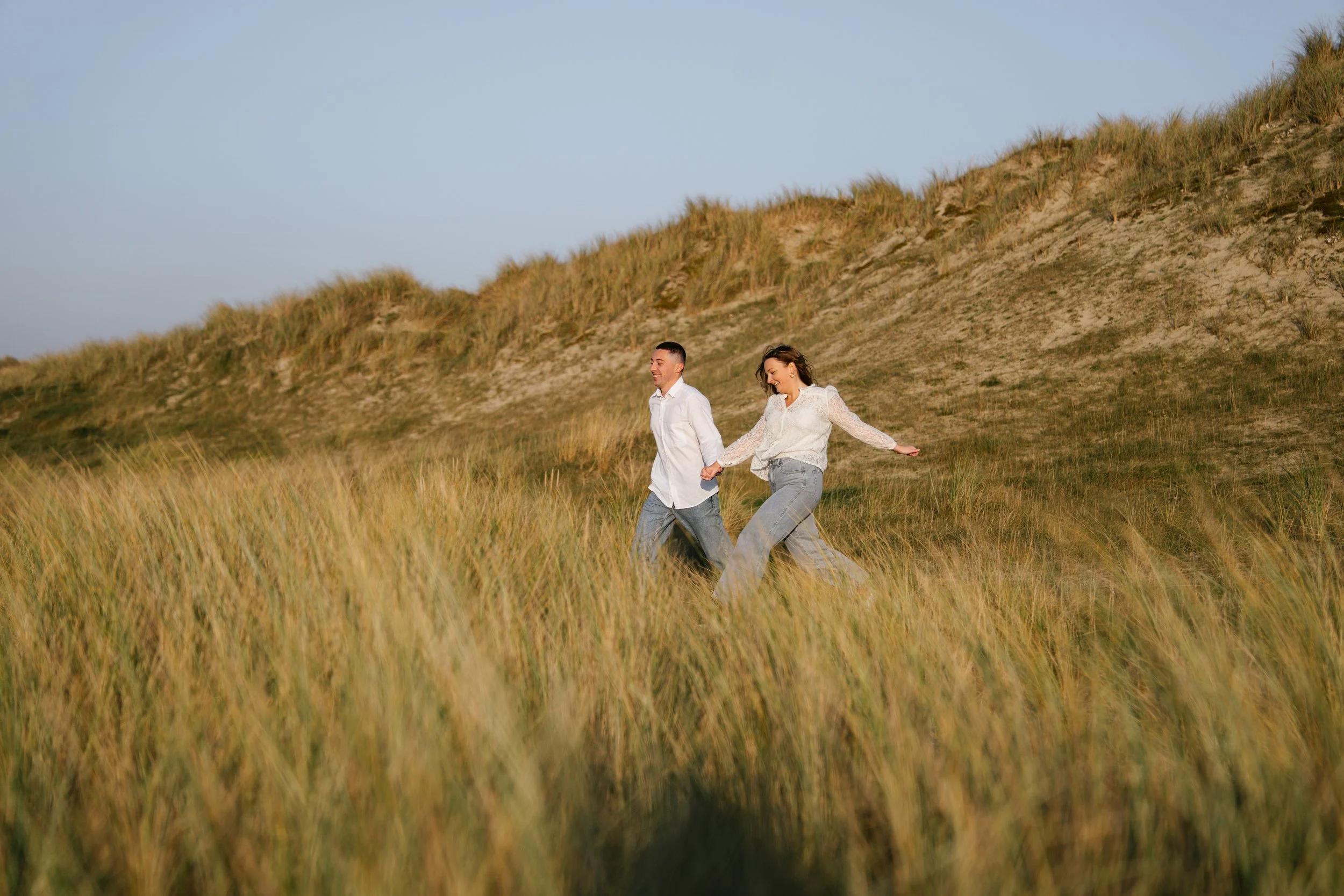 Un couple qui marche dans une prairie d'herbes hautes avec une colline en arrière-plan, lors d'une journée ensoleillée.