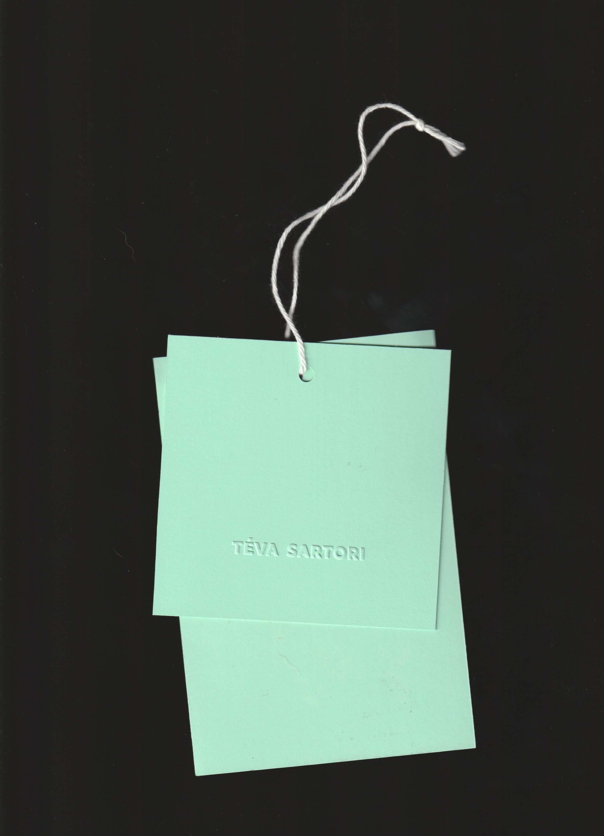 Deux étiquettes en papier de couleur vert menthe avec le nom Tēva Sartori embossé, attachées par un fil blanc sur un fond noir.