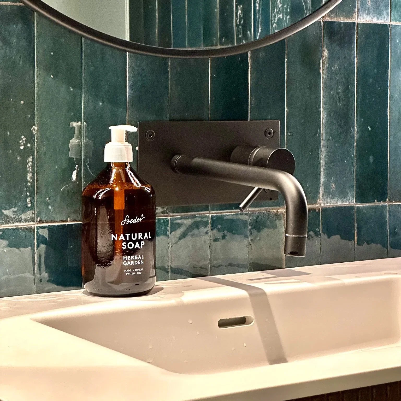 En bademagnet med brunt glasskrukke med såpe, plassert på en vask i et badeværelse med grønn-glaserte fliser. Soeder såper. Soeder Natural care.