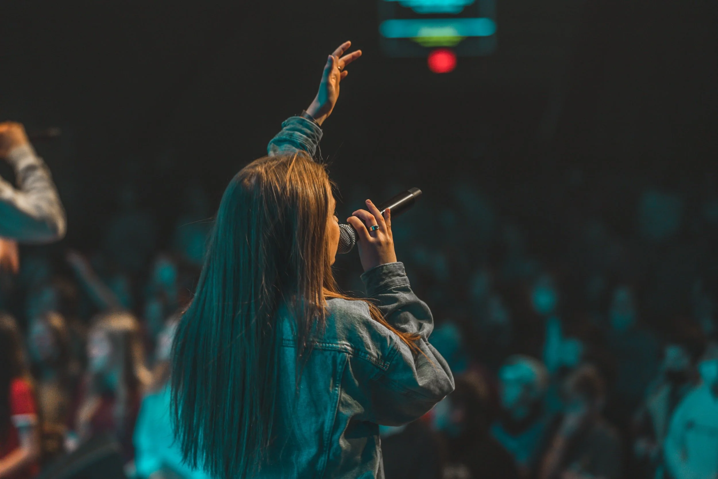 A woman with long brown hair speaking into a microphone while raising her right hand in a dark room with a crowd in the background.