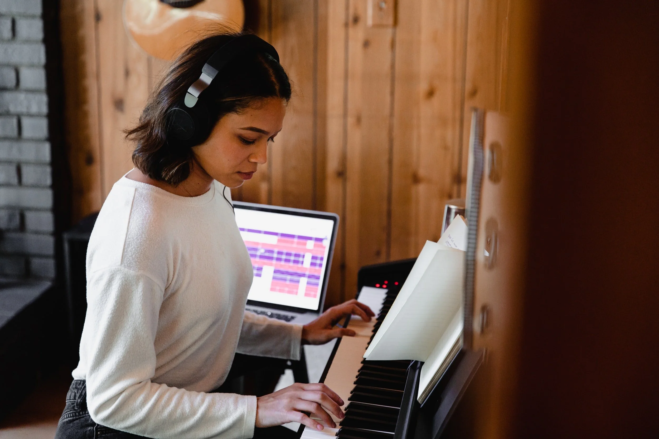 A young woman with dark hair wearing headphones and a white long-sleeve shirt playing a digital piano with sheet music, in a room with wooden walls and a laptop open nearby.
