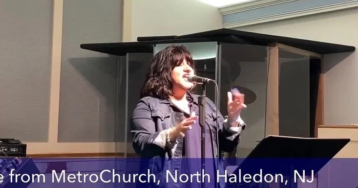 A woman with dark curly hair singing or speaking into a microphone in front of a music stand in a studio or church setting, with a banner that reads 'MetroChurch, North Haledon, NJ' at the bottom.